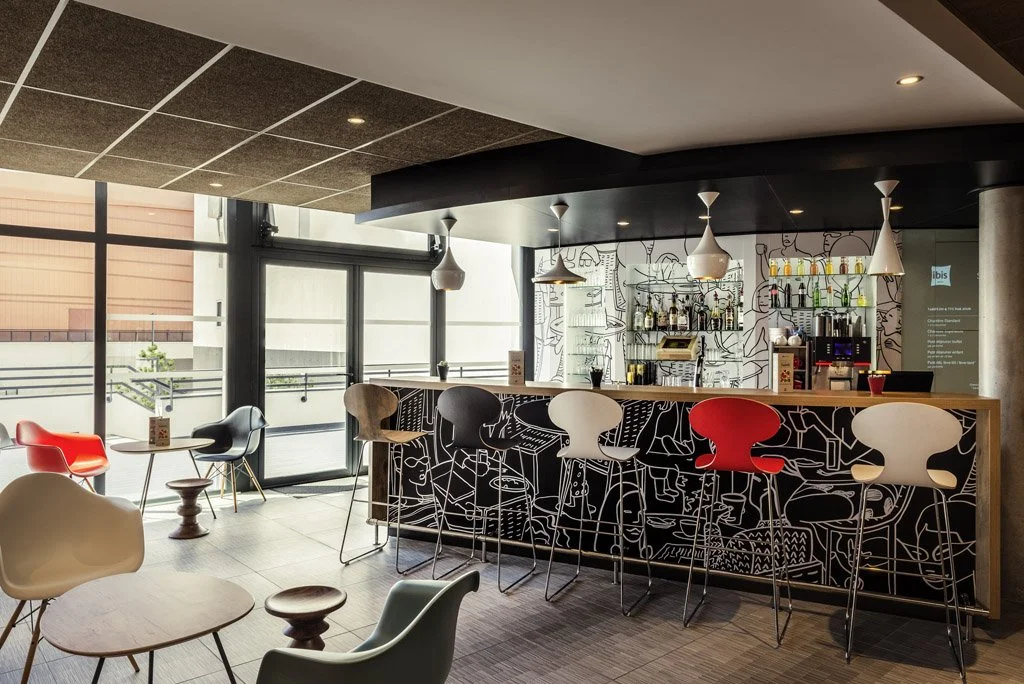 Modern bar with colorful chairs, a small seating area near large windows, and a shelf with bottles behind the counter.
