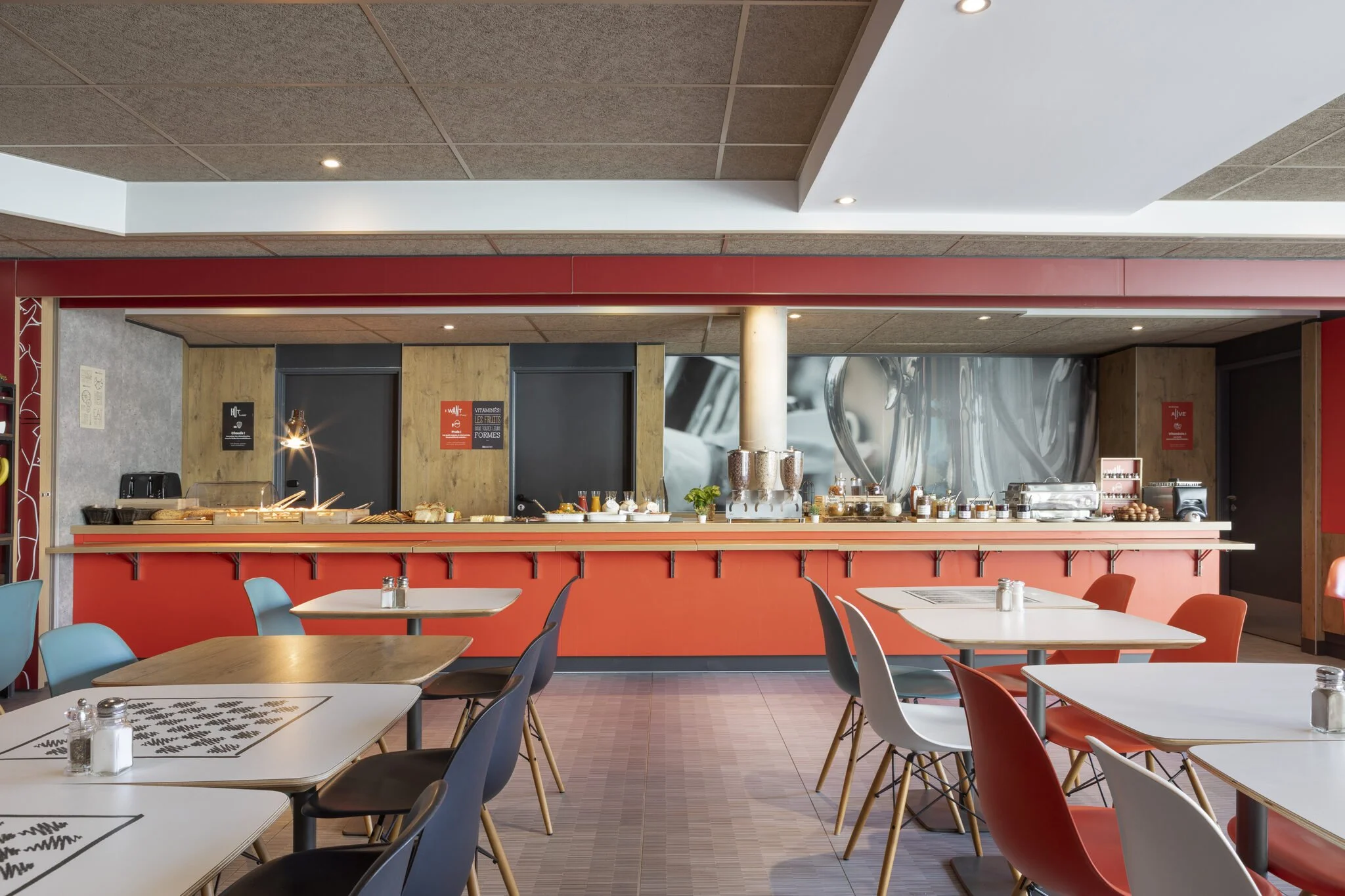 A breakfast buffet in a restaurant with tables and chairs, food and drinks on the counter, and modern decor featuring wooden walls and metal panels.