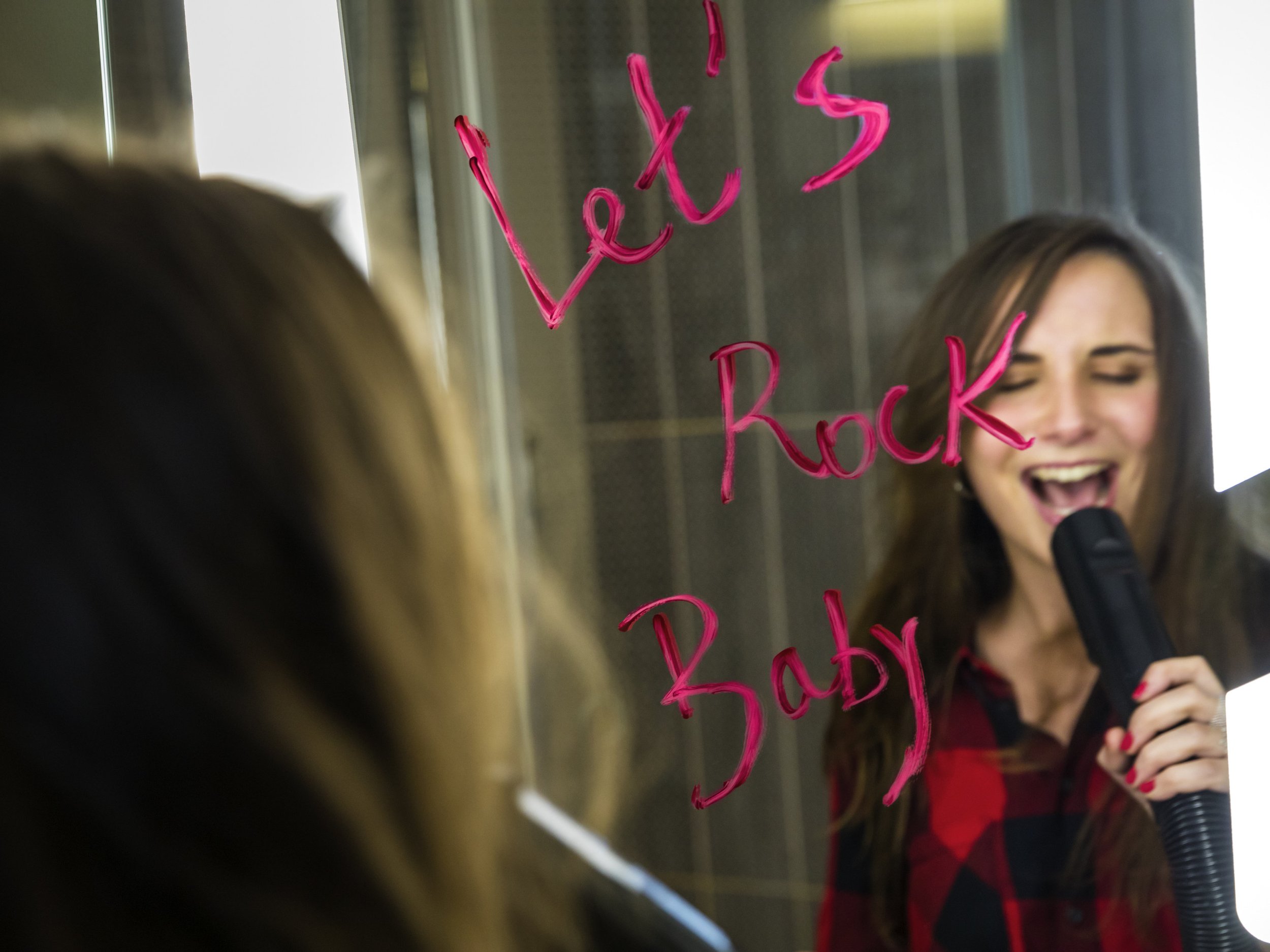 A woman singing or speaking into a microphone, with pink writing on a mirror in front of her that reads 'Let's rock you baby' at Ibis Saint quentin en yvelines.