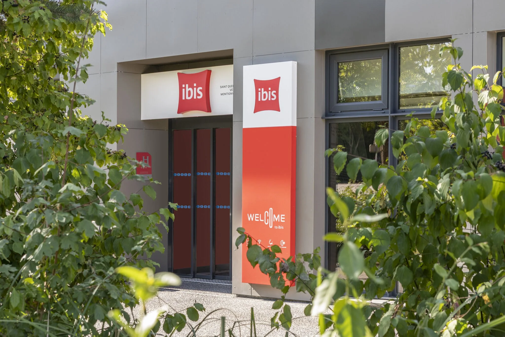 Entrada de un hotel ibis con dos carteles, uno encima y otro al lado, con el logotipo de ibis en rojo sobre fondo blanco. Hay un árbol con hojas verdes que oculta la vista de la entrada.