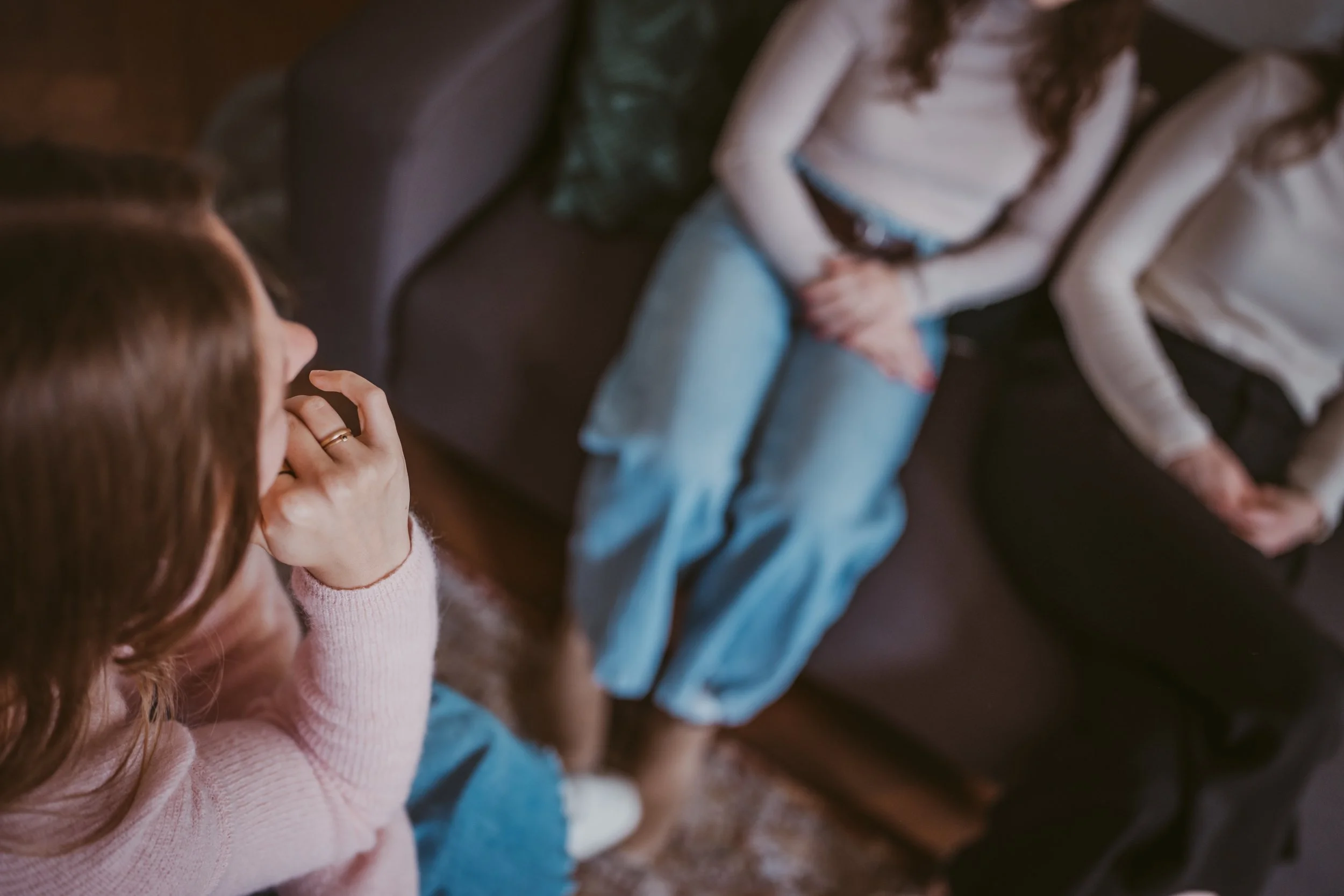 Eine Gruppe von Kindern, die auf einem Sitz in einem Raum sitzen, wobei der Fokus auf einem Mädchen mit lockigem roten Haar liegt, das nachdenklich aussieht. Der Blick ist nach unten gerichtet, und sie trägt einen rosa Pullover.