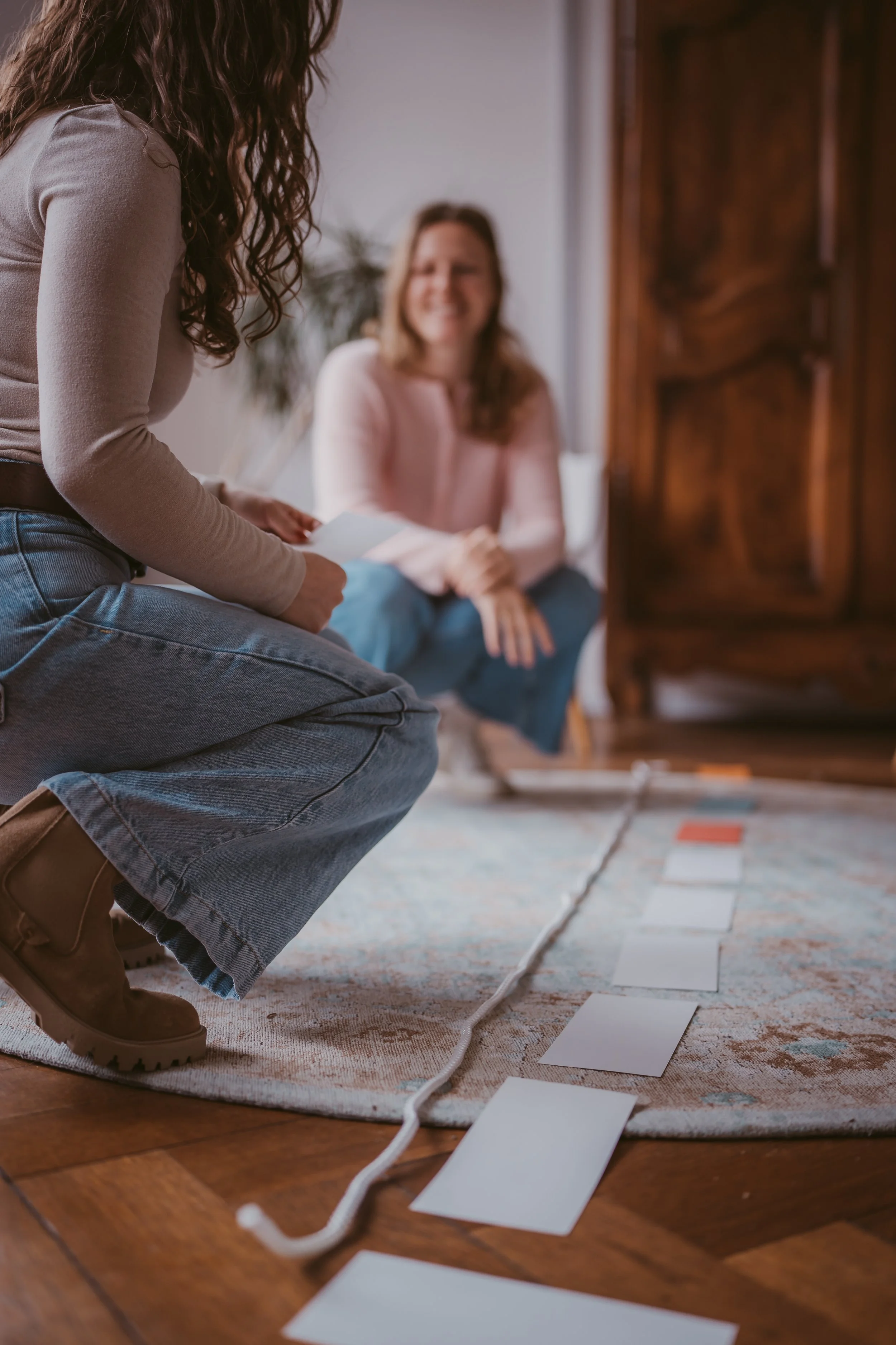 Zwei Frauen sitzen auf einem dicken Teppich im Wohnzimmer, eine arbeitet mit weißen Karten, die andere lacht.