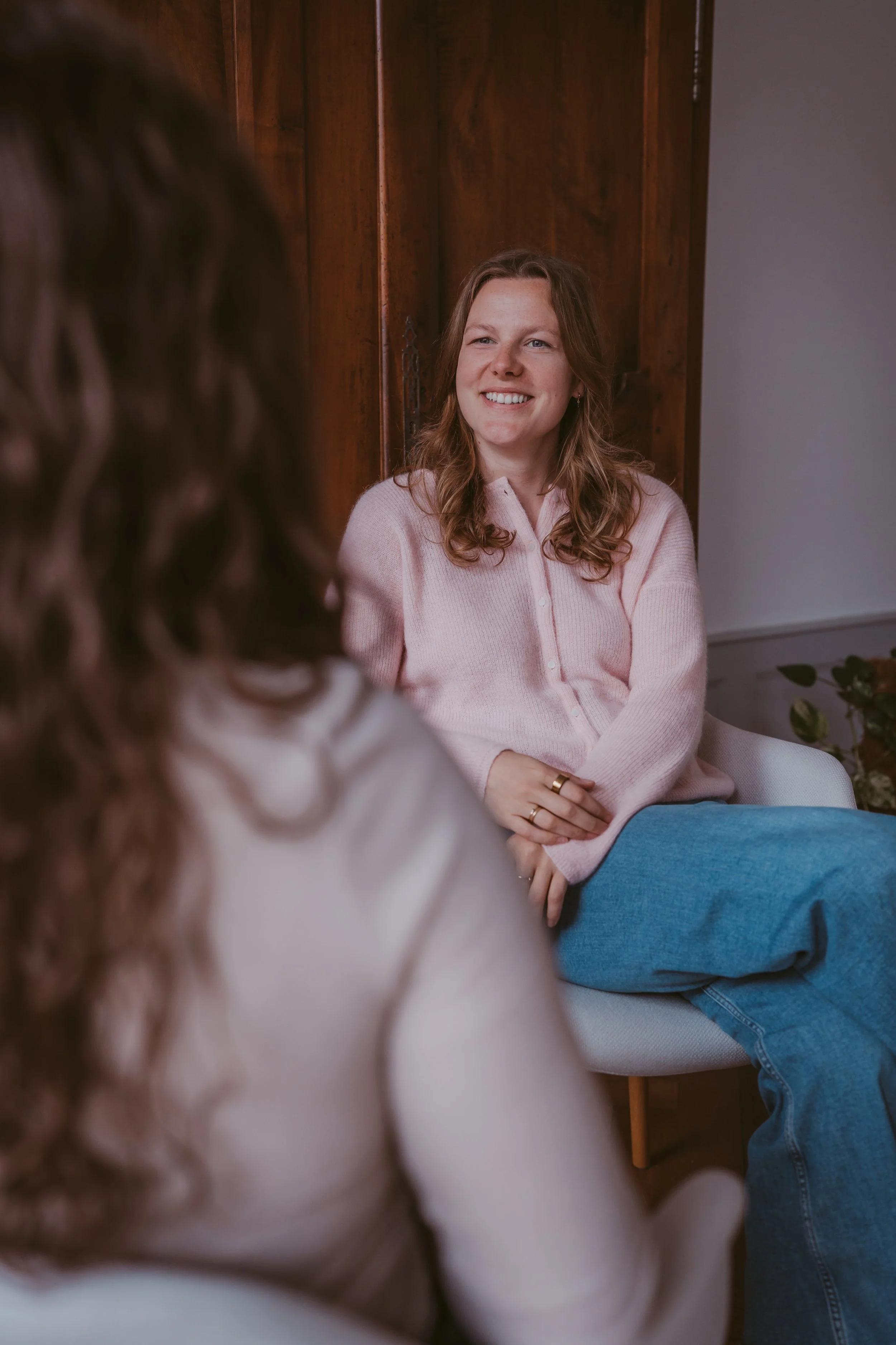 Eine junge Frau mit lockigem braunem Haar sitzt auf einem Sofa, trägt einen hellrosa Pullover, lächelt und sieht in die Kamera, während sie mit jemand anderem spricht, der nur teilweise im Bild zu sehen ist.