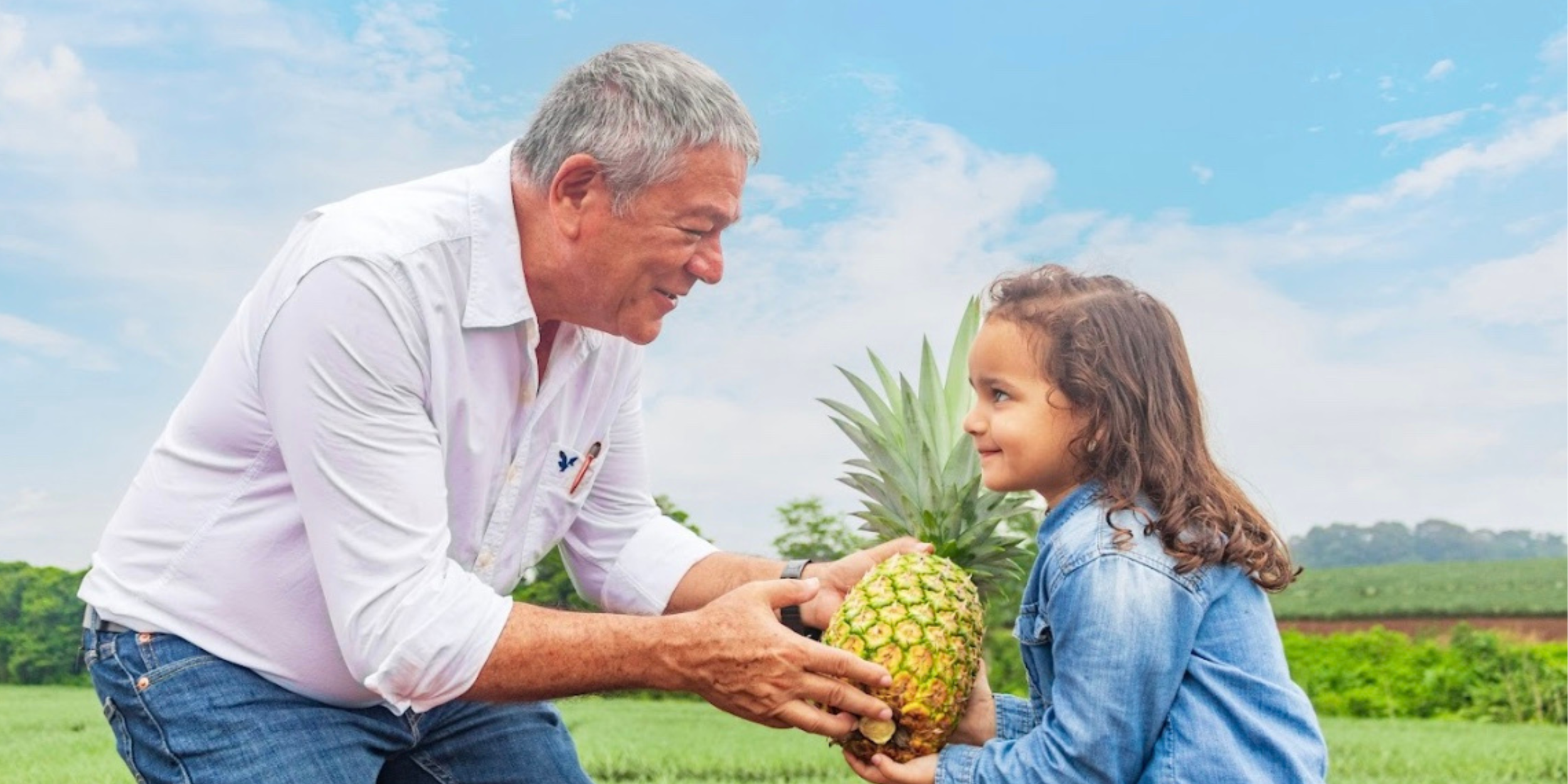 An elderly man and a young girl exchange a pineapple outdoors in a green field under a blue sky with scattered clouds. The man is smiling and wearing a white shirt, and the girl is wearing a denim jacket, looking at the man.