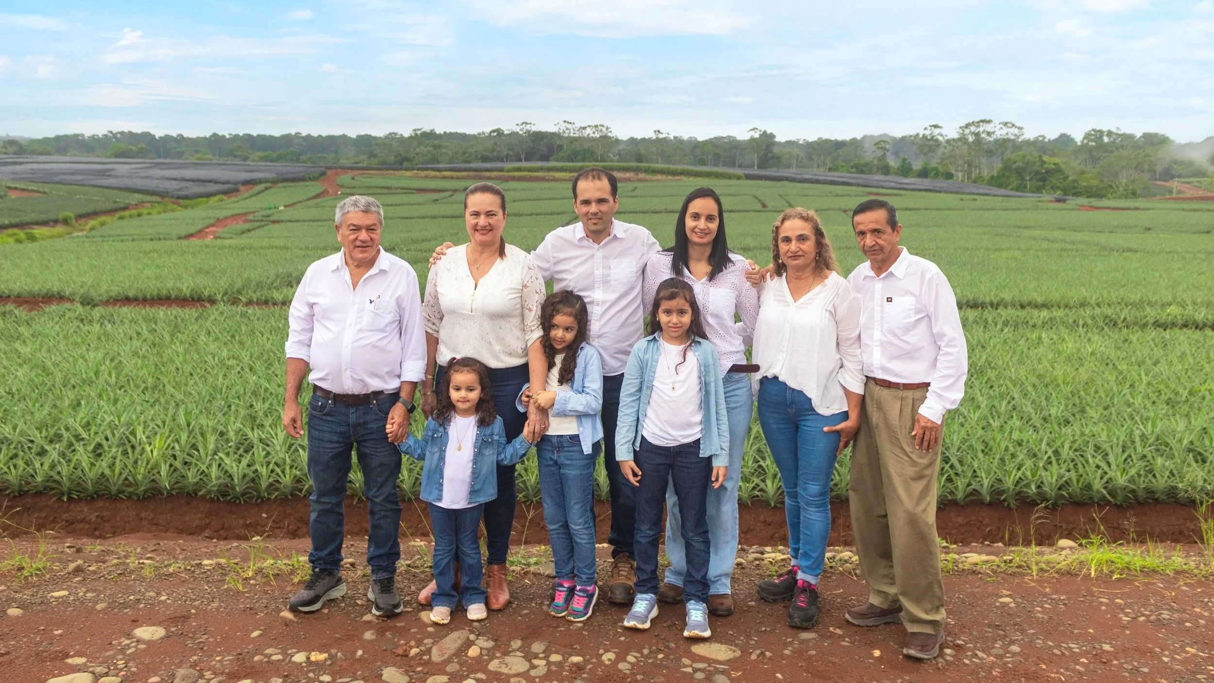 Group of nine people, including children, standing in front of a green agricultural field with cloudy sky in the background.