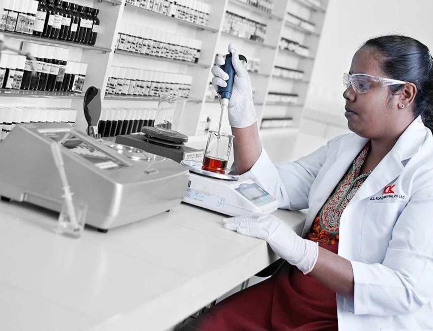 A scientist in a laboratory wearing safety goggles and gloves, using a pipette to transfer a red liquid into a test tube.