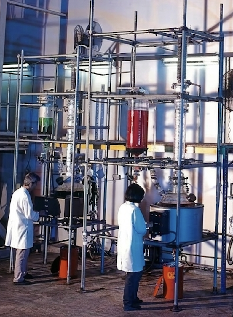Scientists in lab coats examining a large complex industrial setup with pipes, tanks, and gauges, likely conducting a chemical or scientific experiment.