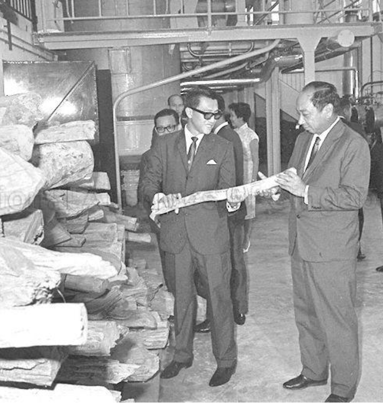 Two men in suits and glasses examine a large, rolled-up item in an industrial or warehouse setting, with shelves and other people in the background.