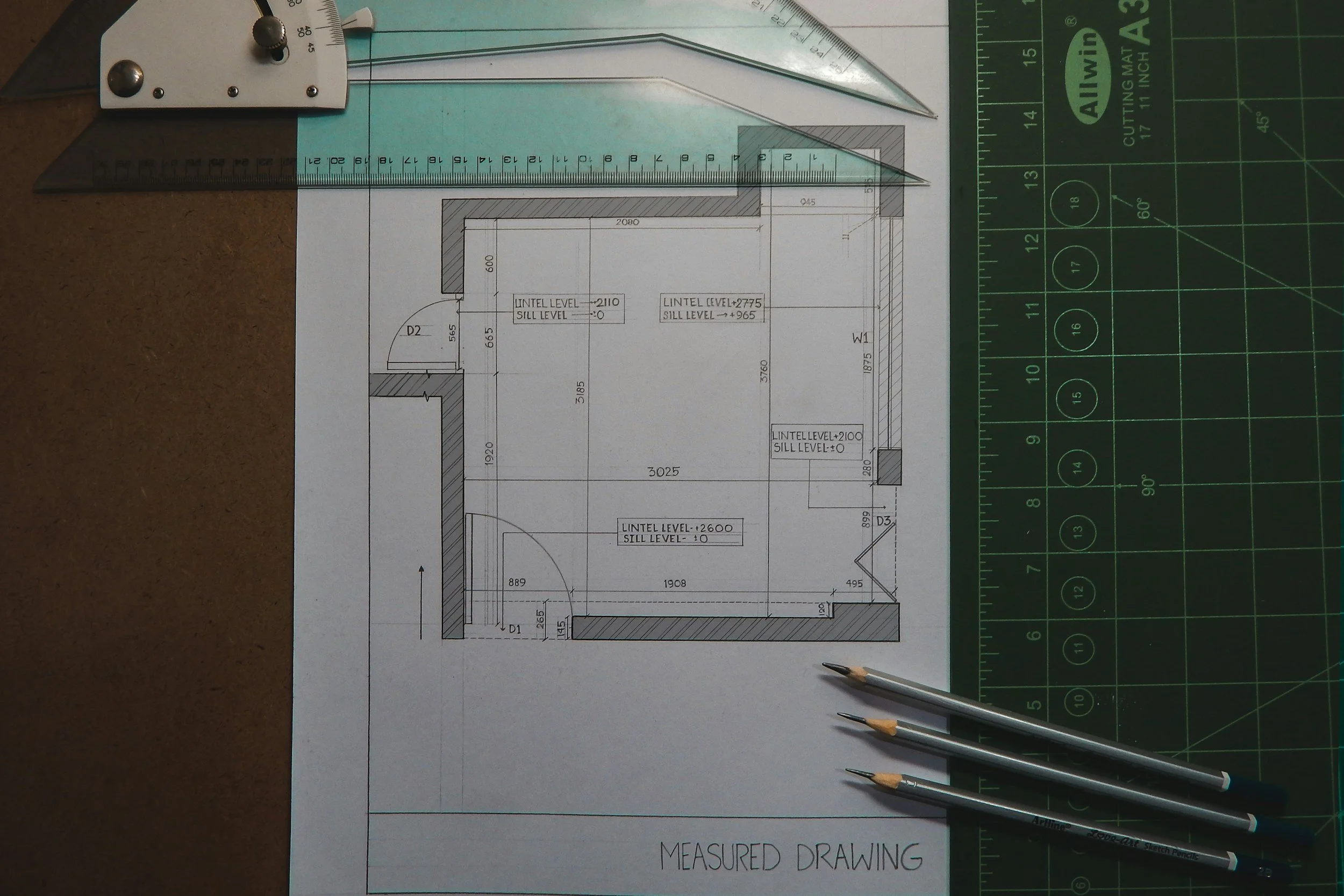 Architectural blueprint of a room layout with various measurements and labels, placed on a work surface with drafting tools including a metal ruler, triangle, and three pencils.
