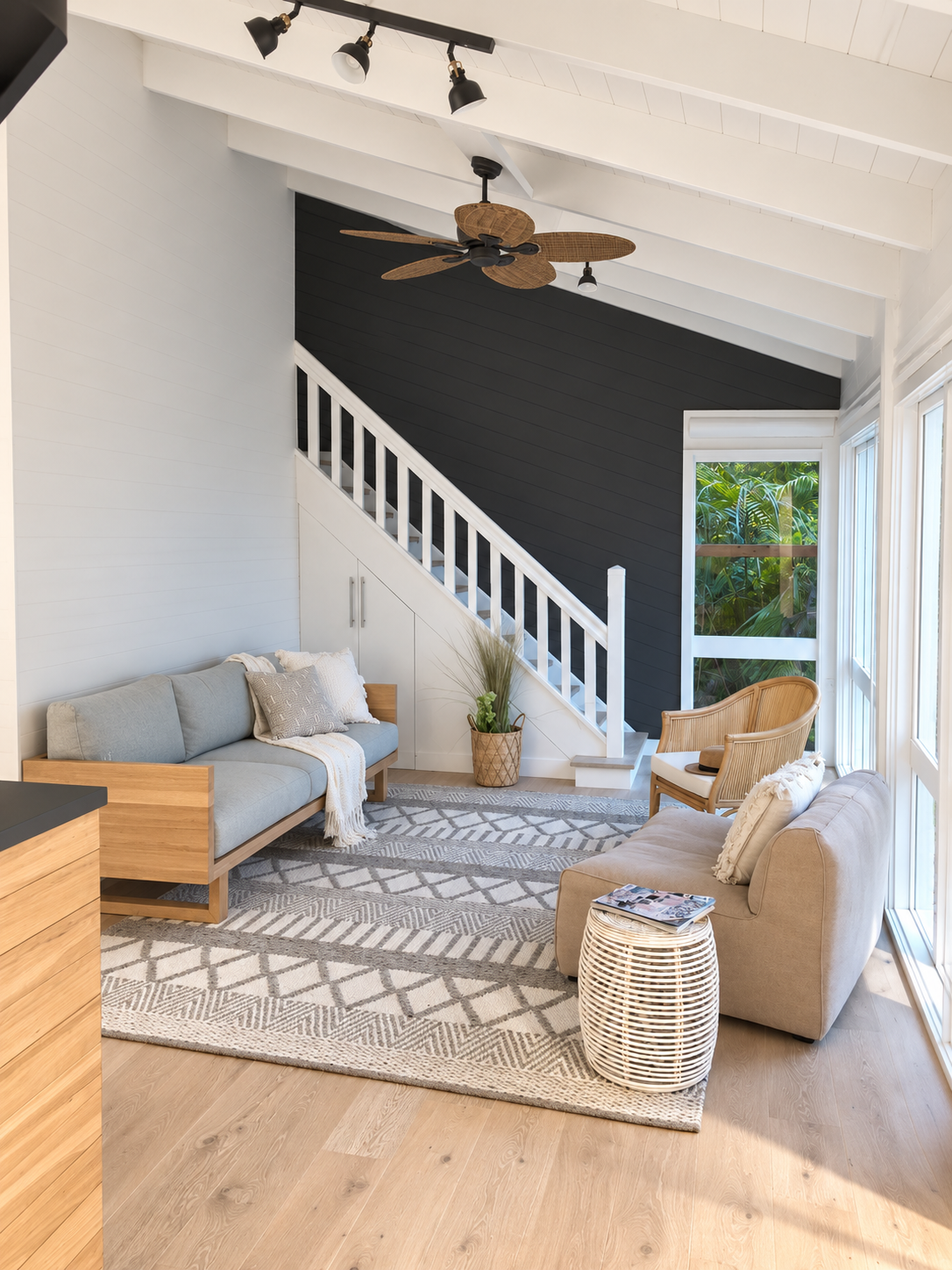 Living room with beige and gray furniture, a patterned area rug, wooden accents, a staircase with a dark wall behind it, and large windows showing greenery outside.