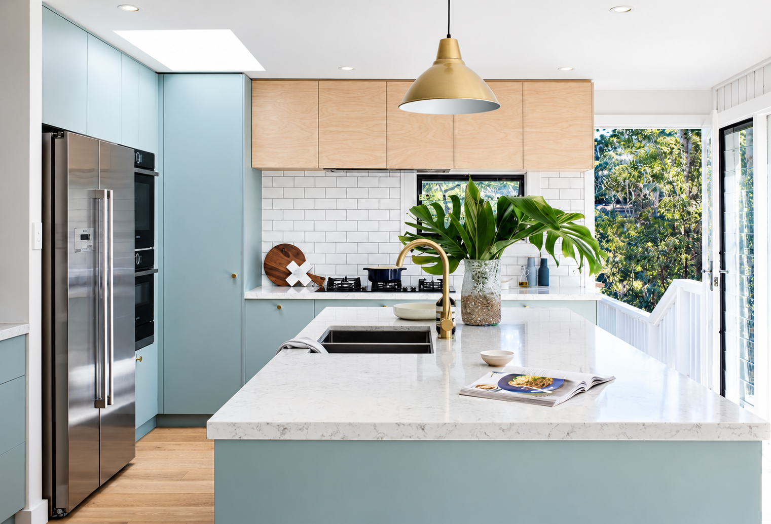 Modern kitchen with blue cabinets, white marble countertops, and a large green plant on the island. Stainless steel refrigerator, white subway tile backsplash, wooden upper cabinets, and large window with a view of trees.