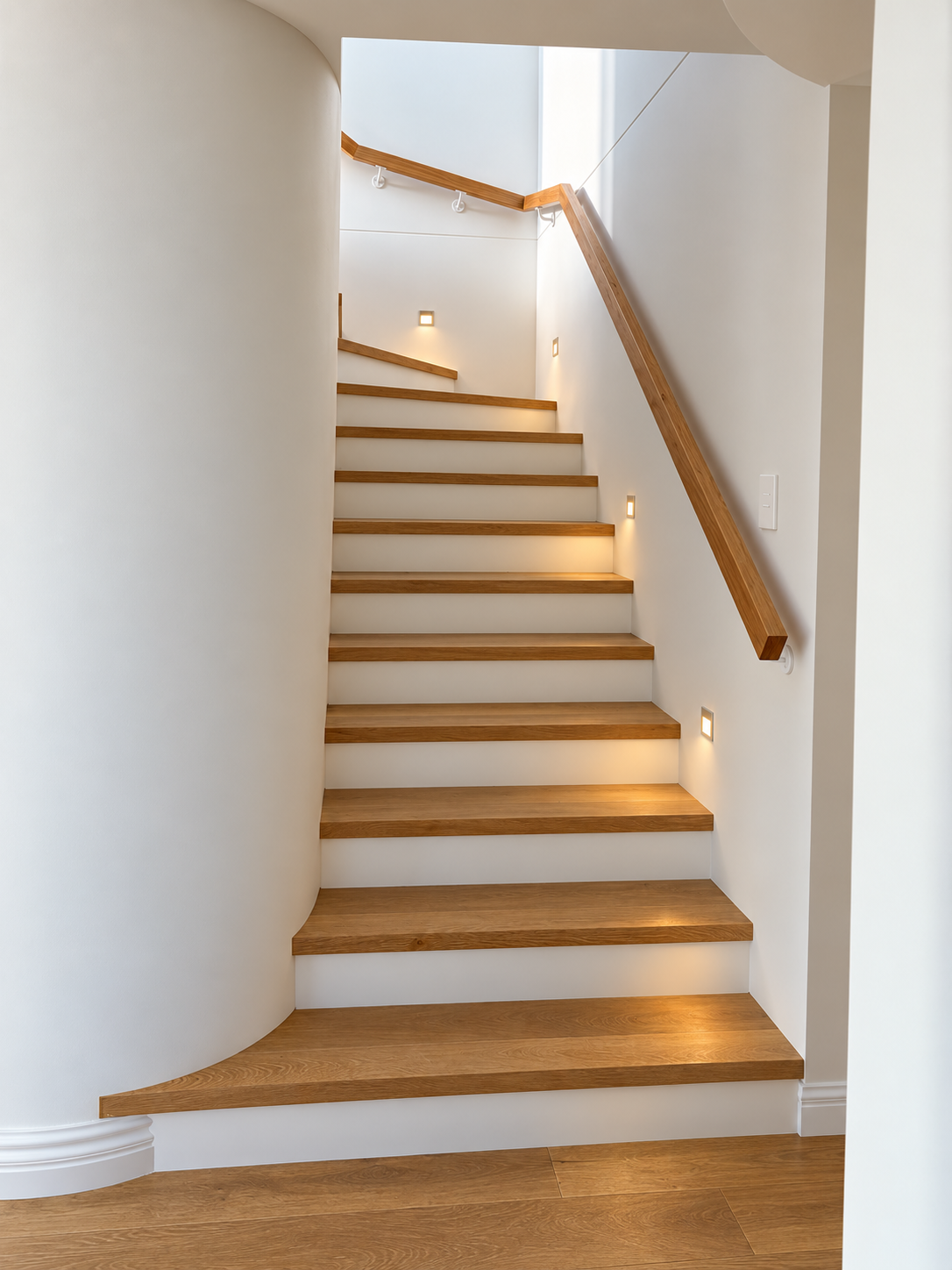 Modern wooden staircase with integrated lighting, curved white wall, and minimalistic design in a bright, contemporary home.