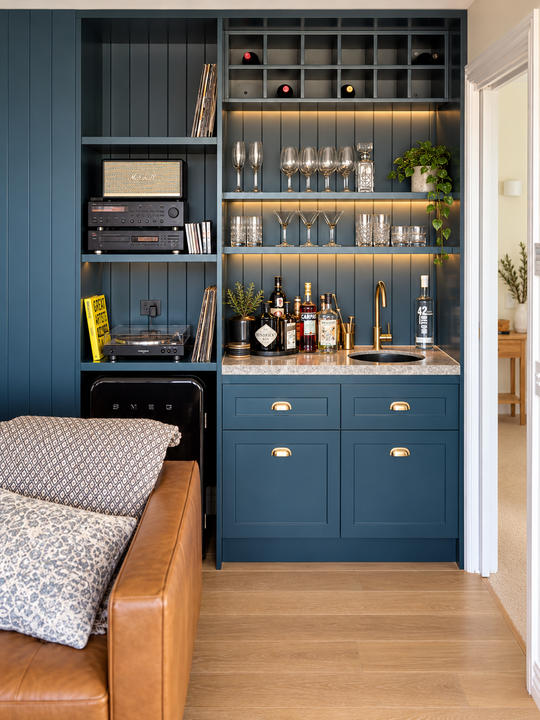A modern living space with a built-in bar area featuring blue cabinetry, wine bottles, glassware, and liquor bottles. There are shelves with glasses and decorative items, a small sink with a gold faucet, and a countertop. To the left, part of a brown