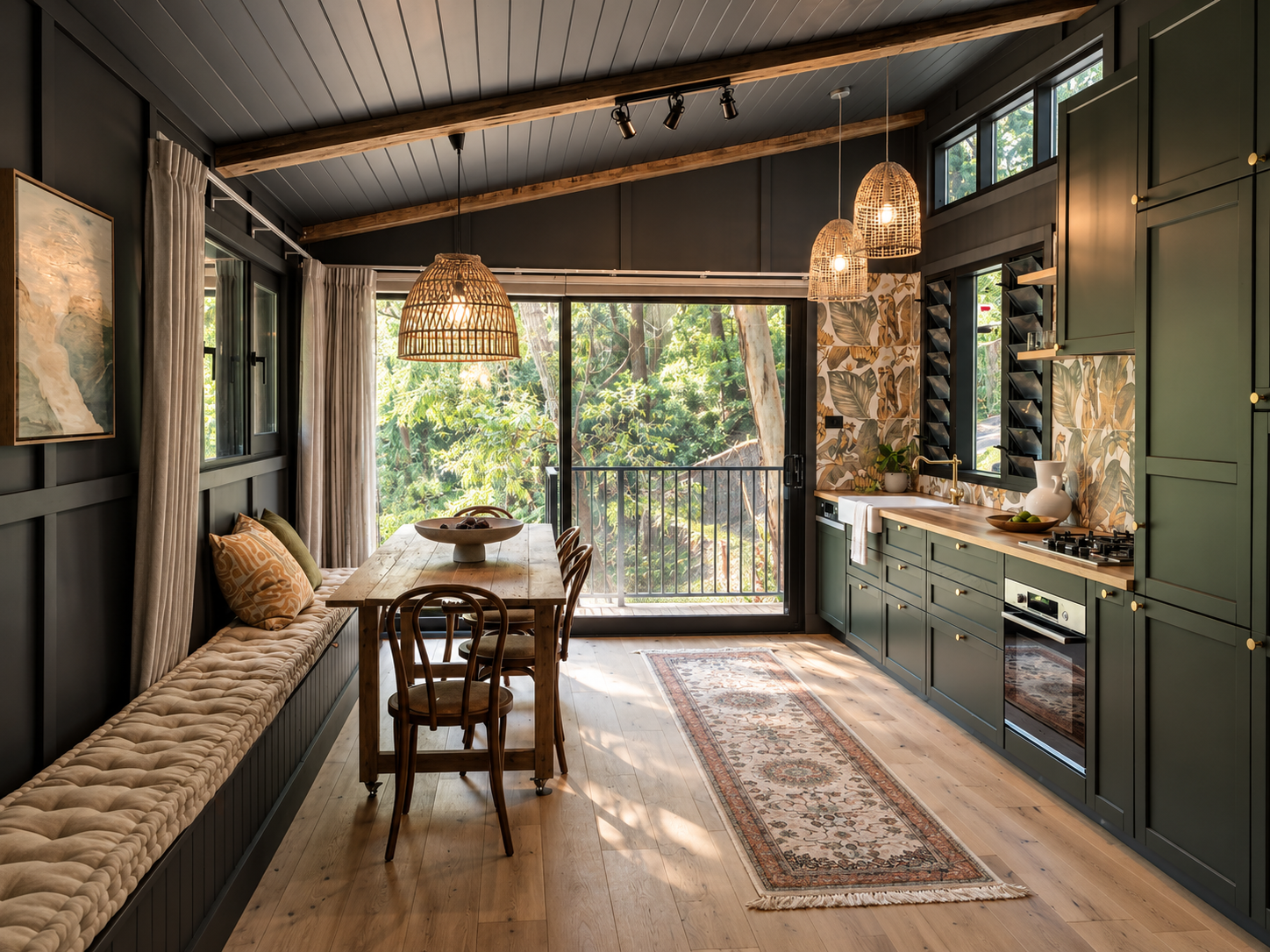 A cozy kitchen and dining area with green cabinetry, a wooden dining table with chairs, a built-in bench with cushions, large windows, and hanging pendant lights, overlooking lush outdoor greenery.