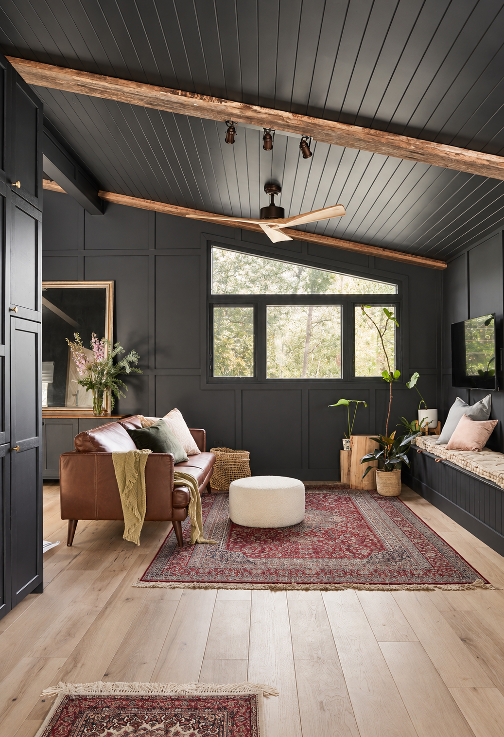 A cozy living room with black paneled walls and ceiling, a brown leather sofa with cushions, a white pouf, potted plants, a large window with trees outside, and a built-in window seat with cushions. There is a red patterned rug on light wood flooring