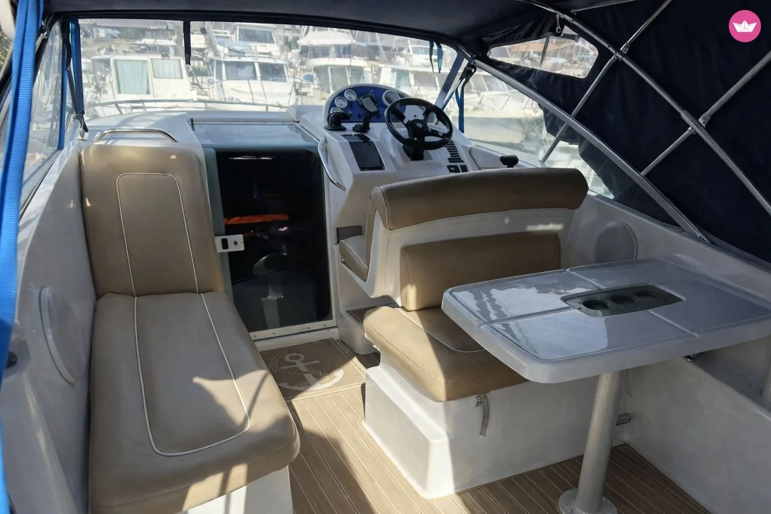Intérieur d'un yacht avec des sièges en cuir beige, un tableau de bord avec plusieurs instruments et un volant, une table pliante blanche avec un compartiment, et une vue de la marina en arrière-plan.Location, bateau, plaisance, cote bleue, carry