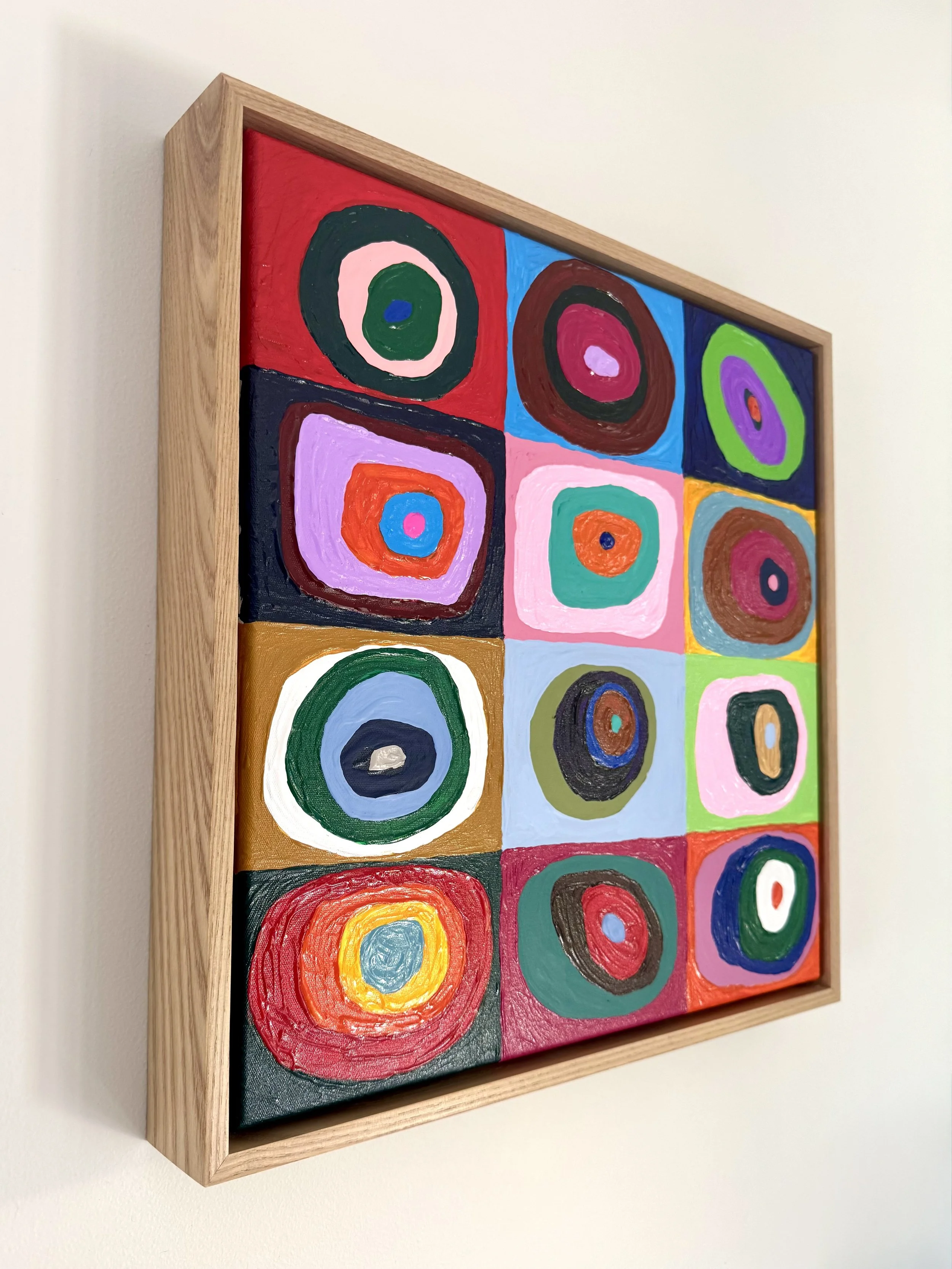 Colorful abstract painting of concentric circles arranged in a grid, framed in light wood, hanging on a white wall.