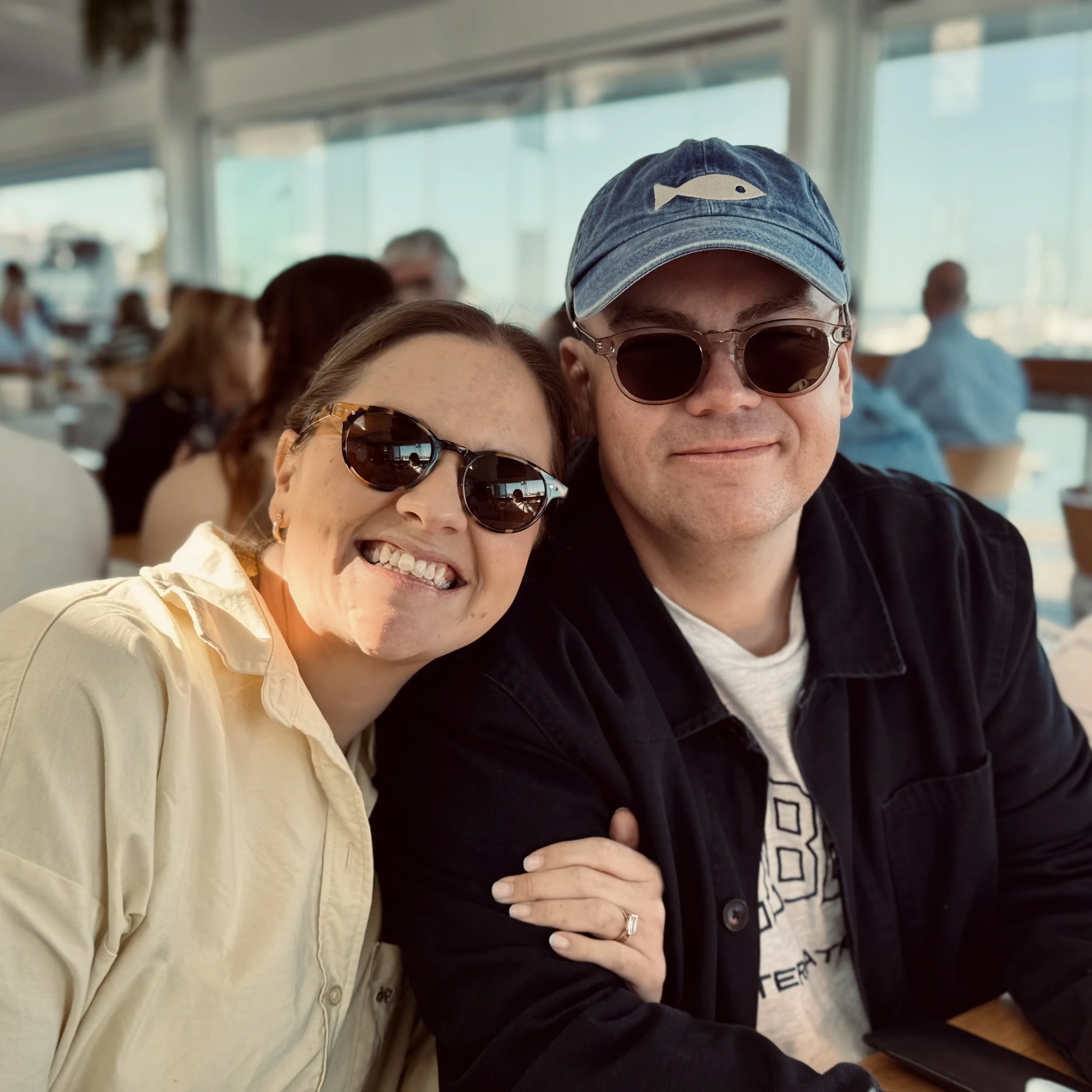 Husband and wife artists sitting closely together at a café, smiling and wearing sunglasses, enjoying a relaxed moment.