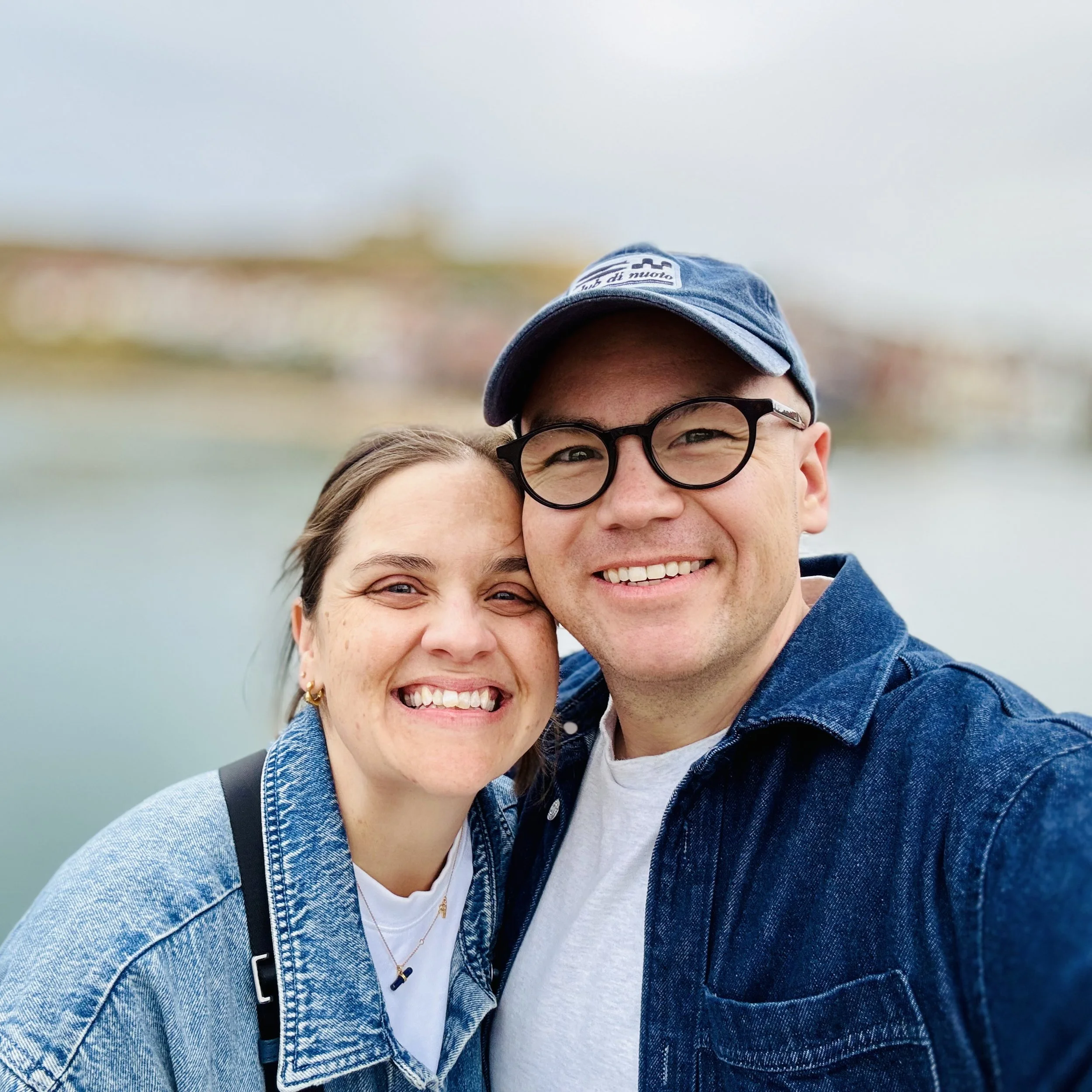 Close-up of a smiling woman and a smiling man, with a blurred background and the man wearing a blue cap and glasses.
