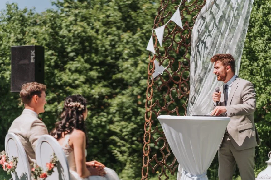 Ein Brautpaar sitzt im Freien, während ein Mann im Anzug eine Rede hält, während einer Hochzeit.