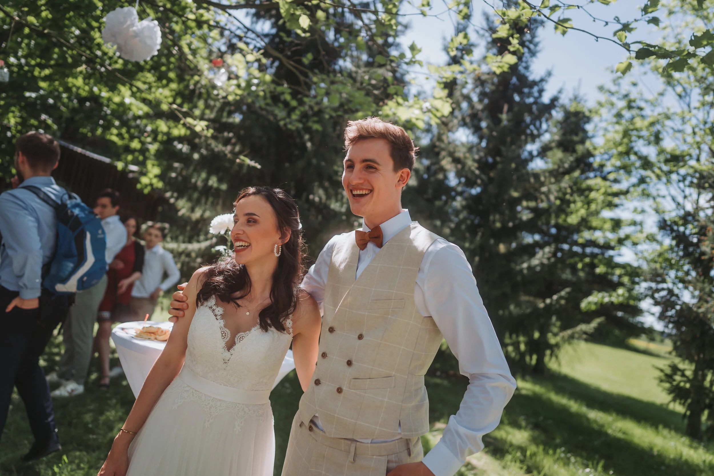 Ein lachendes Brautpaar bei einer Hochzeit im Freien, umgeben von Gästen, unter grünen Bäumen bei sonnigem Wetter.