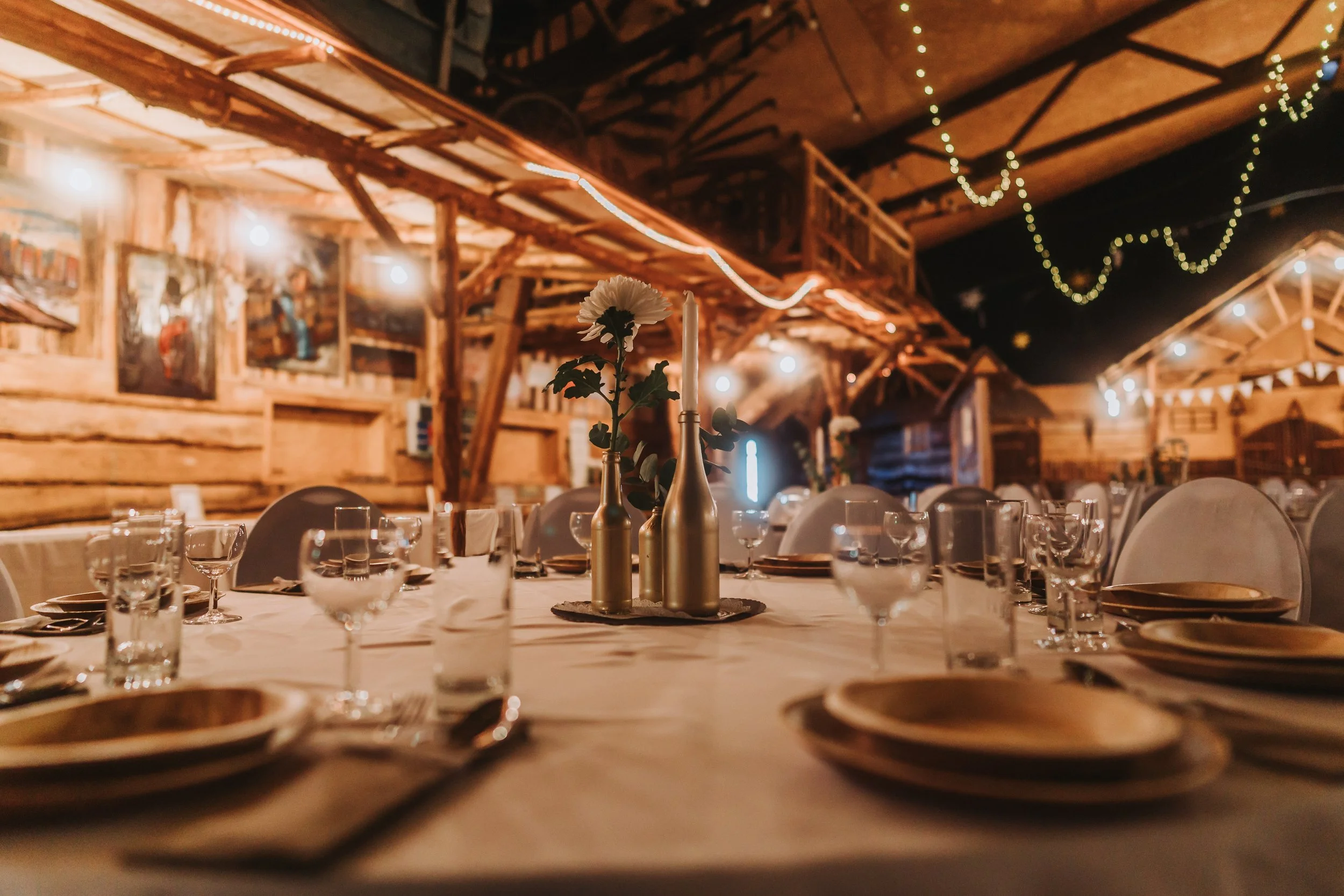 Ein festlich gedeckter Tisch in einer rustikalen Holzhalle mit Kerzen, Blumen und Gläsern, Dekorationen mit Lichterketten im Hintergrund.