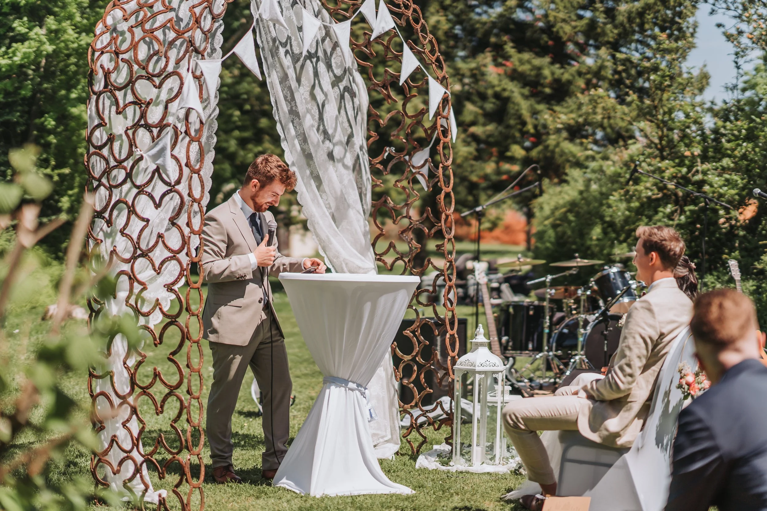 Ein Mann hält eine Rede bei einer Hochzeit im Freien, während andere Gäste den Moment beobachten. Im Hintergrund sind Musikinstrumente und ein Baum zu sehen.
