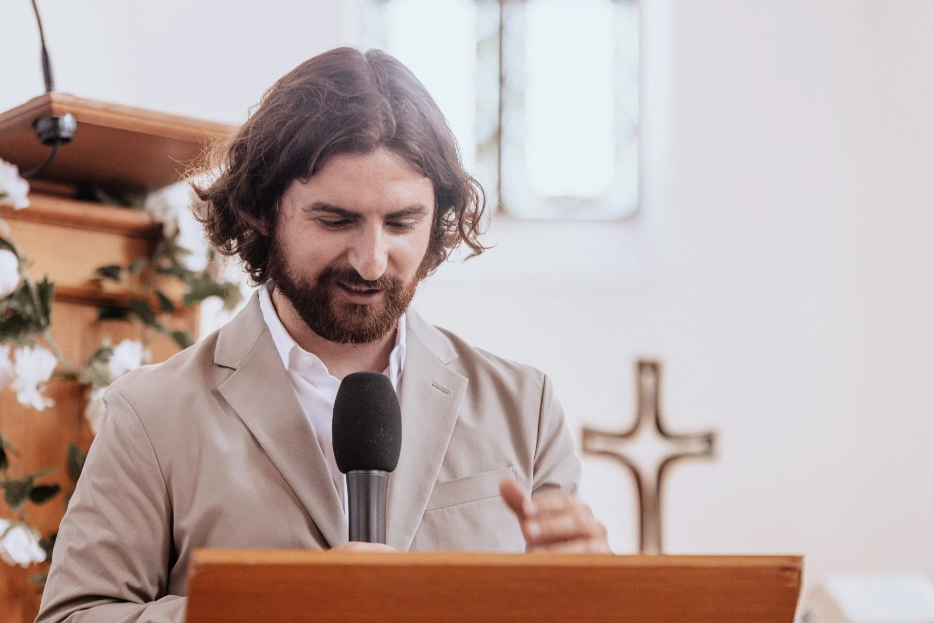 Ein Mann mit braunem, lockigem Haar und Bart, der an einer Predigt in einer Kirche spricht, während er einen schwarzen Mikrofon hält, mit einem Kreuz im Hintergrund.