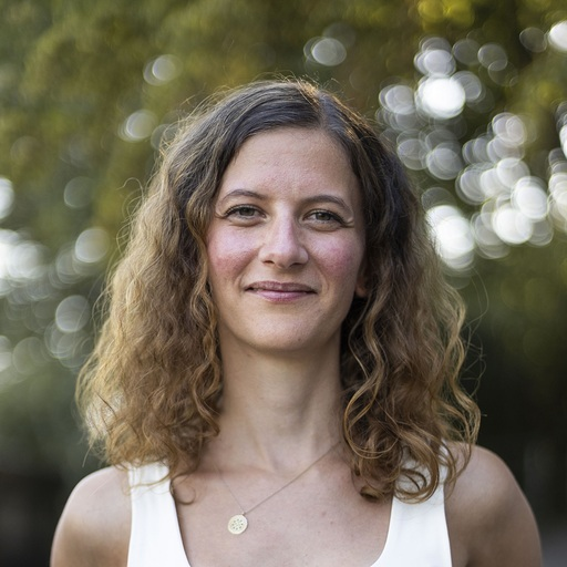 A woman with long, curly brown hair and fair skin outdoors with a blurred background of trees and sunlight.