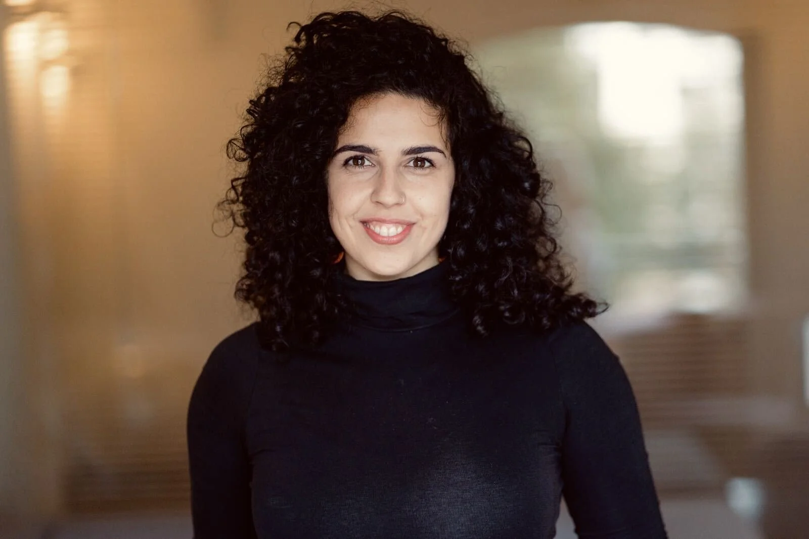 A woman with dark, curly hair, wearing a black turtleneck, smiling with visible teeth, standing in a warmly lit indoor space with blurred background.