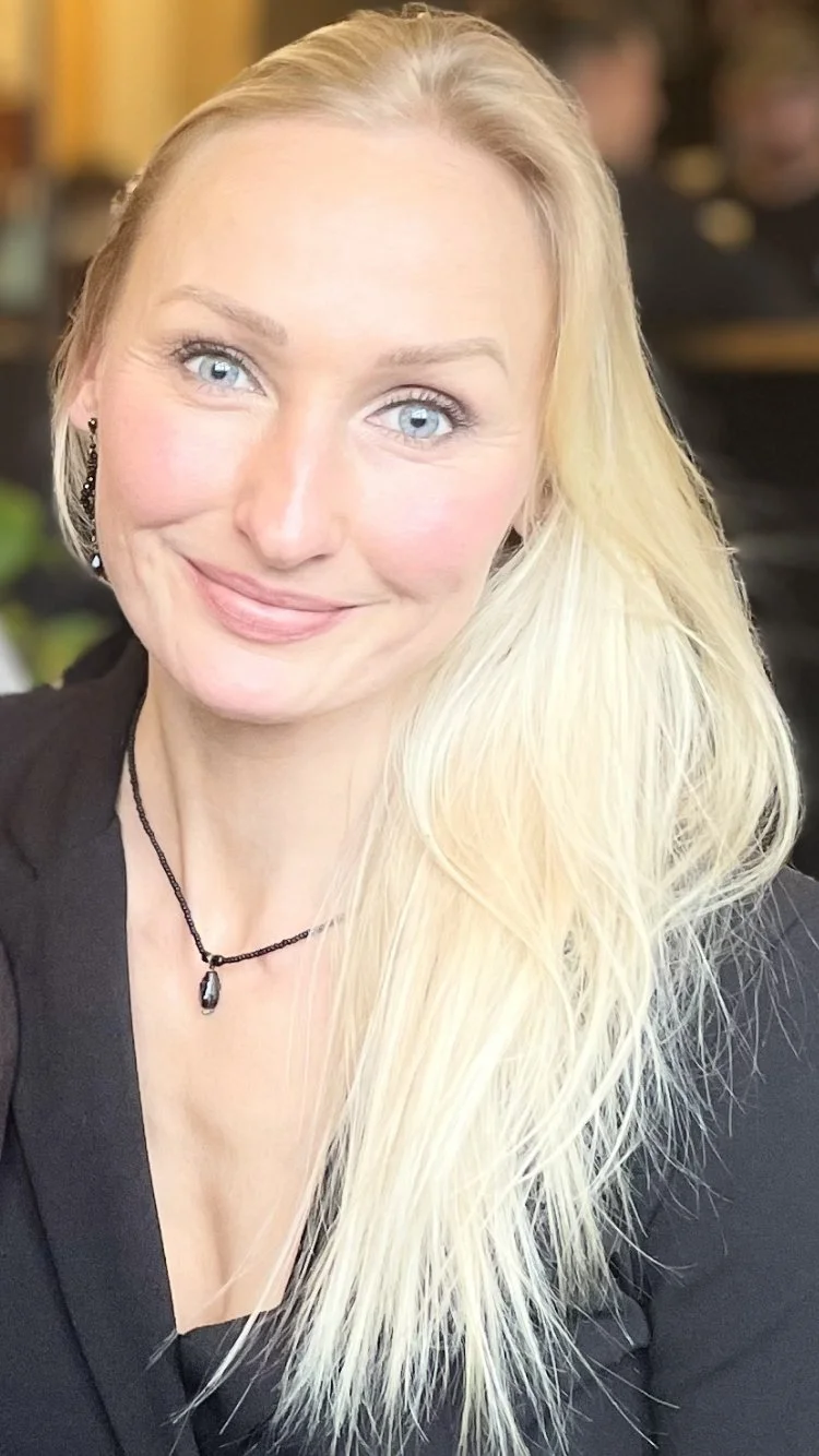 A woman with long blonde hair, blue eyes, and light skin smiling at the camera, wearing a black top, black earrings, and a black necklace with a pendant.