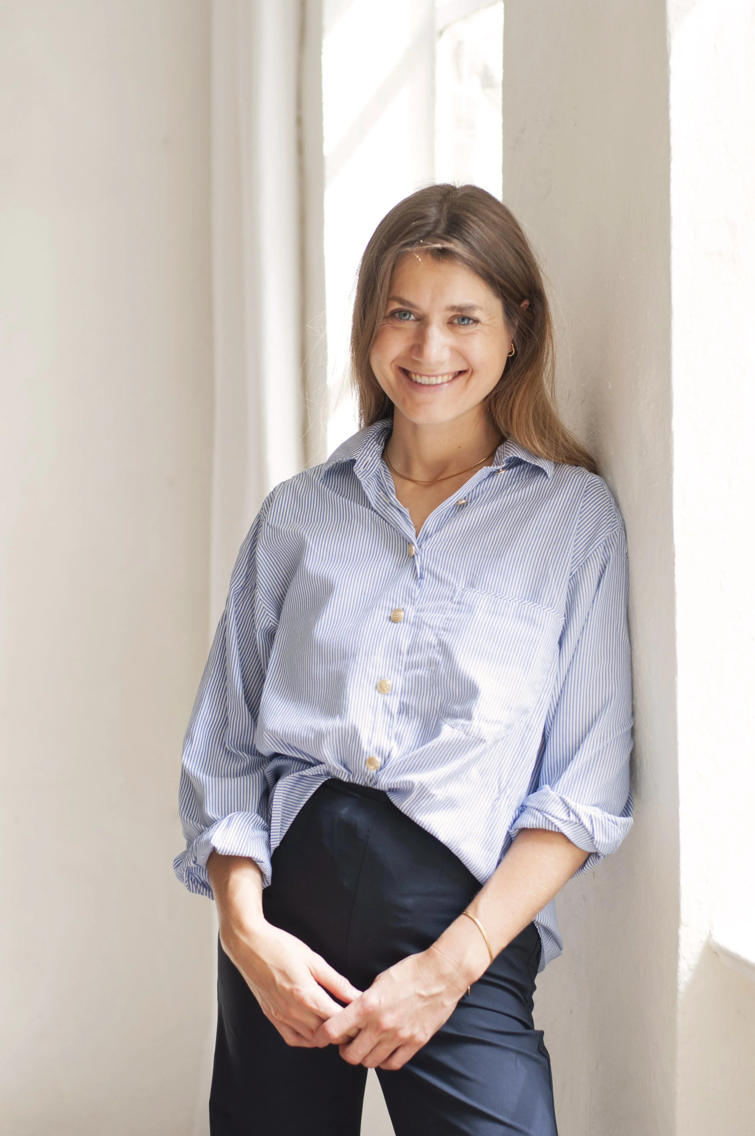 A woman with light brown hair styled in loose waves, wearing a light blue and white striped button-up shirt with the sleeves rolled up and black pants, smiling while standing against a white wall, with sunlight coming through a window in the background.
