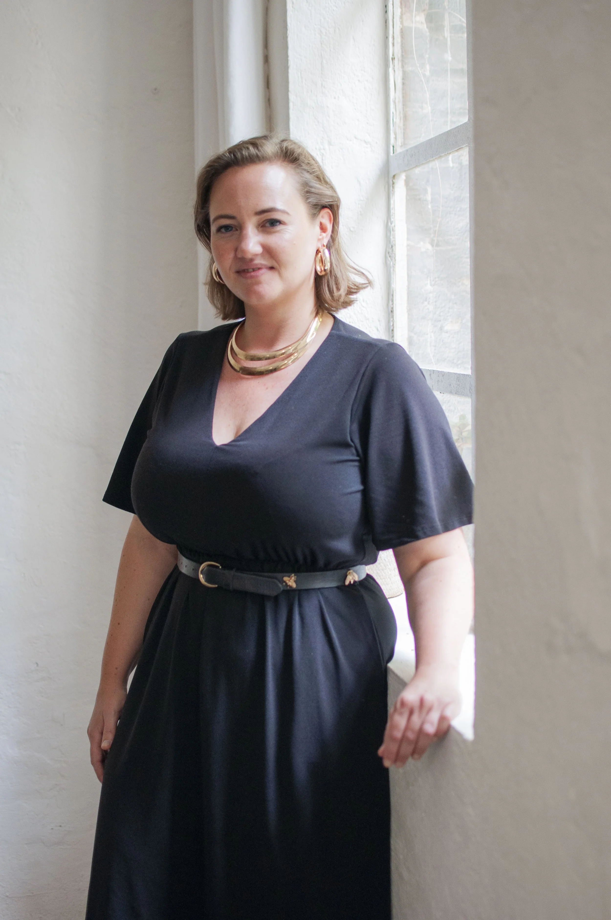 A woman with shoulder-length brown hair, wearing a black dress with a smile, standing near a window with a white wall background.