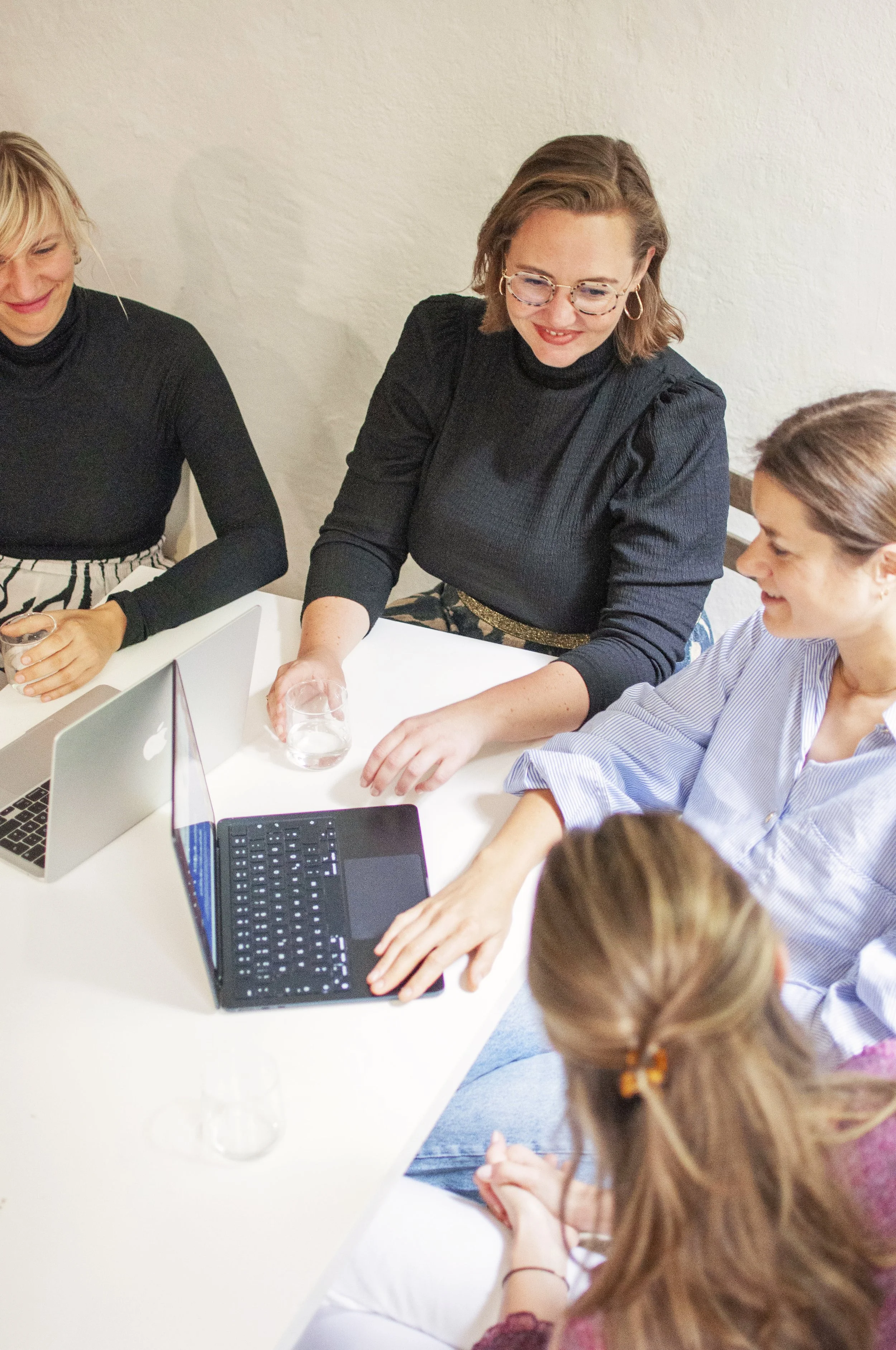 Four women are sitting around a white table, working together with laptops and papers. They appear engaged and smiling in a meeting or collaborative work session.