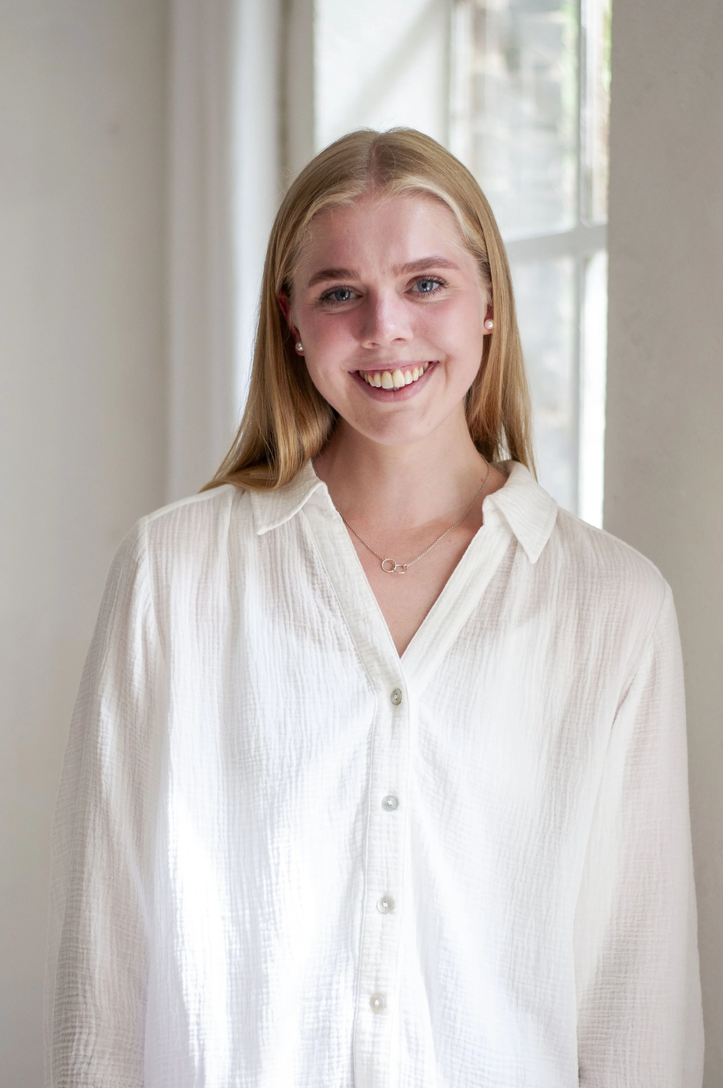 A woman with blonde hair, blue eyes, and light skin, wearing a white button-up shirt with pearl earrings and a silver necklace, standing indoors near a window with sunlight streaming in.