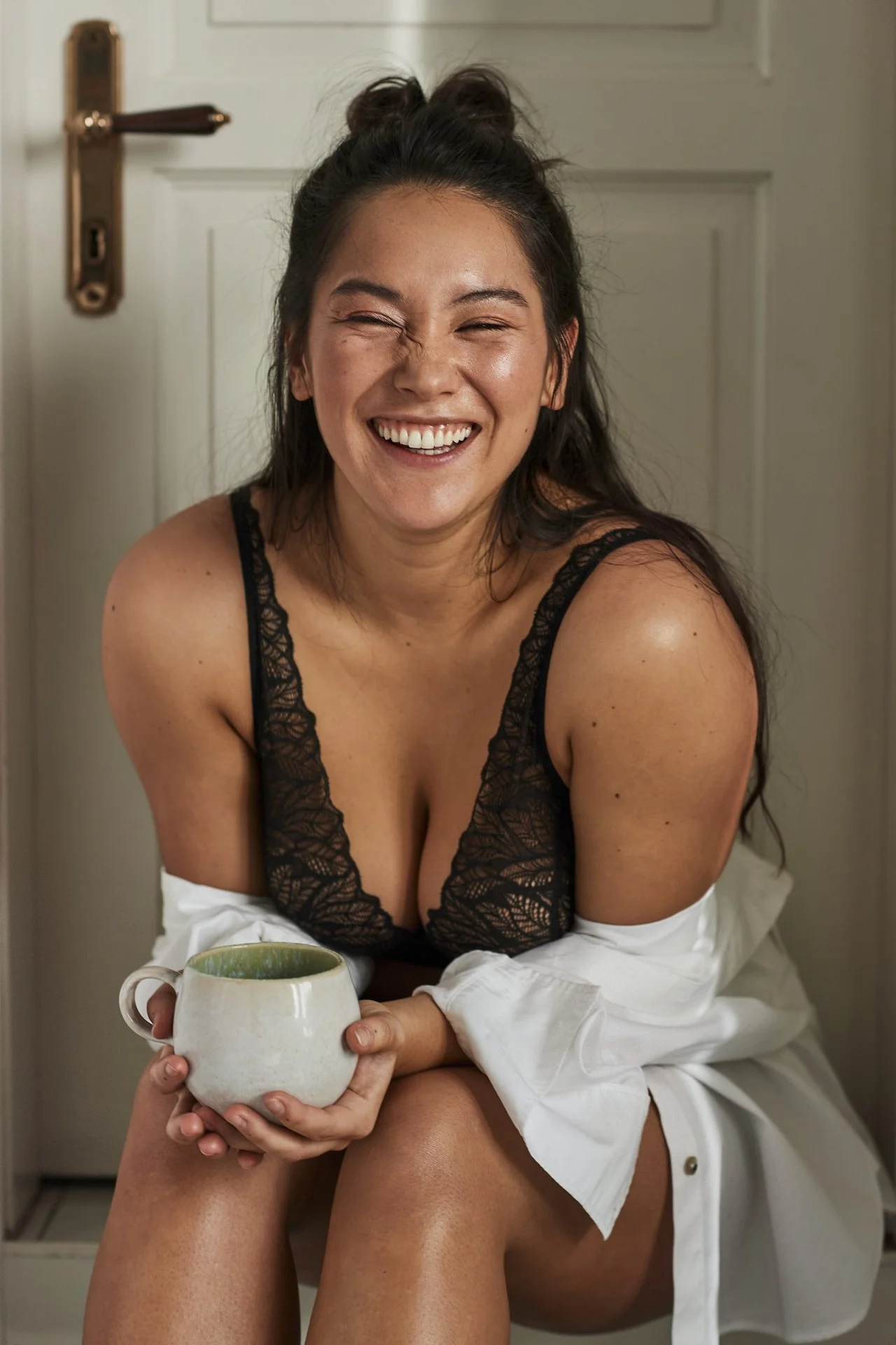 Eine lachende Frau mit dunklen Haaren, trägt einen schwarzen Spitzenbrusthalter und hält eine weiße Tasse. Sie sitzt in einem Raum vor einer weißen Tür.