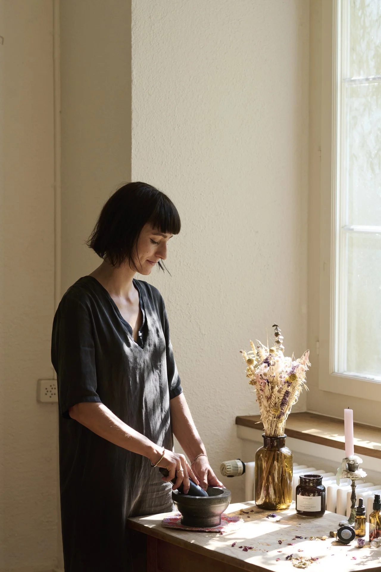Eine Frau mit dunklen Haaren, die einen schwarzen Mörser und Stößel in einem hellen Raum benutzt, mit Sonnenlicht, das durch ein Fenster fällt, auf einem Tisch mit Blumen, Kerzen und Flaschen