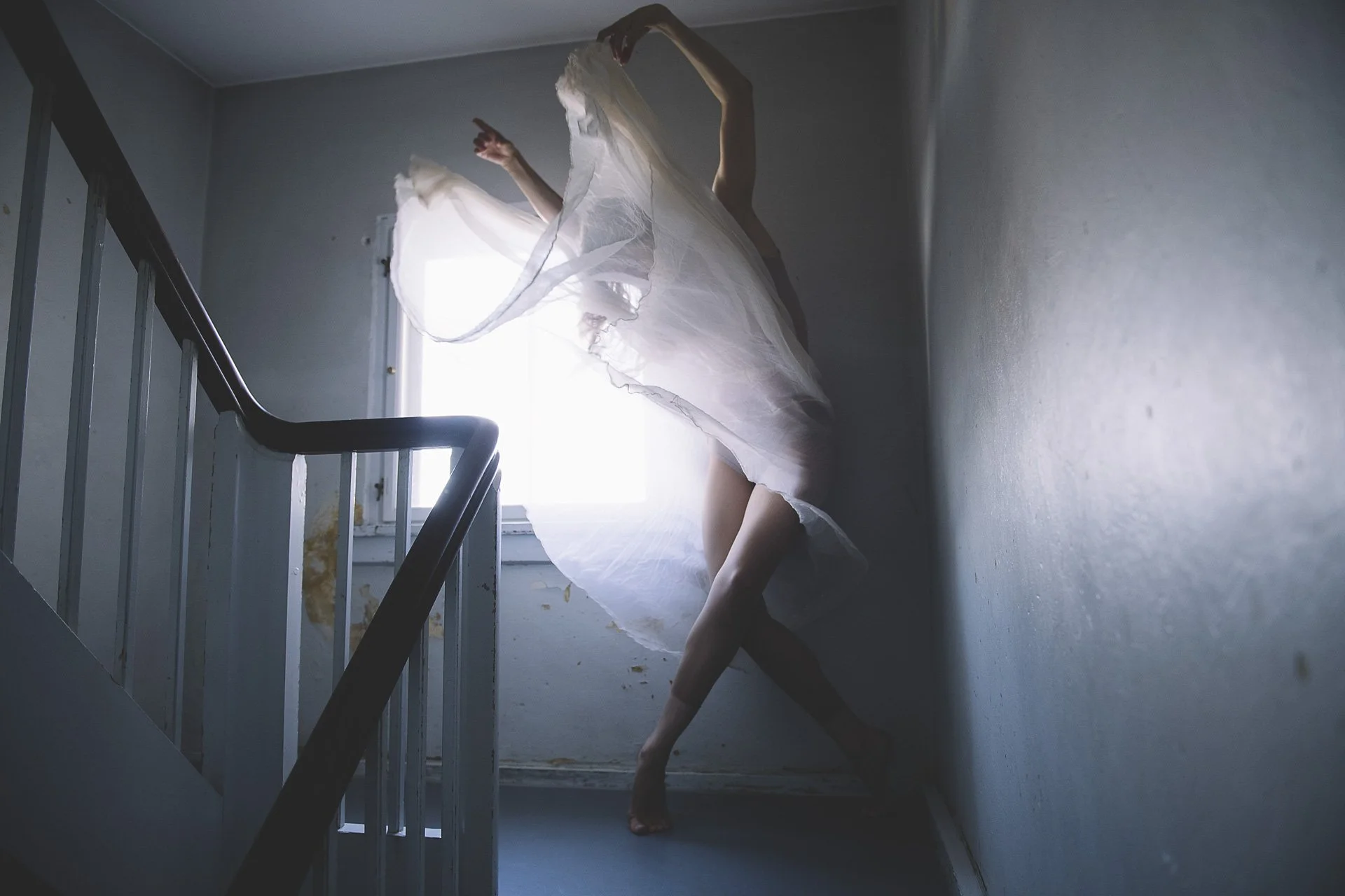 Tänzerin in einem weißen Kleid tanzt in einem dunklen, engen Flur bei Tageslicht.