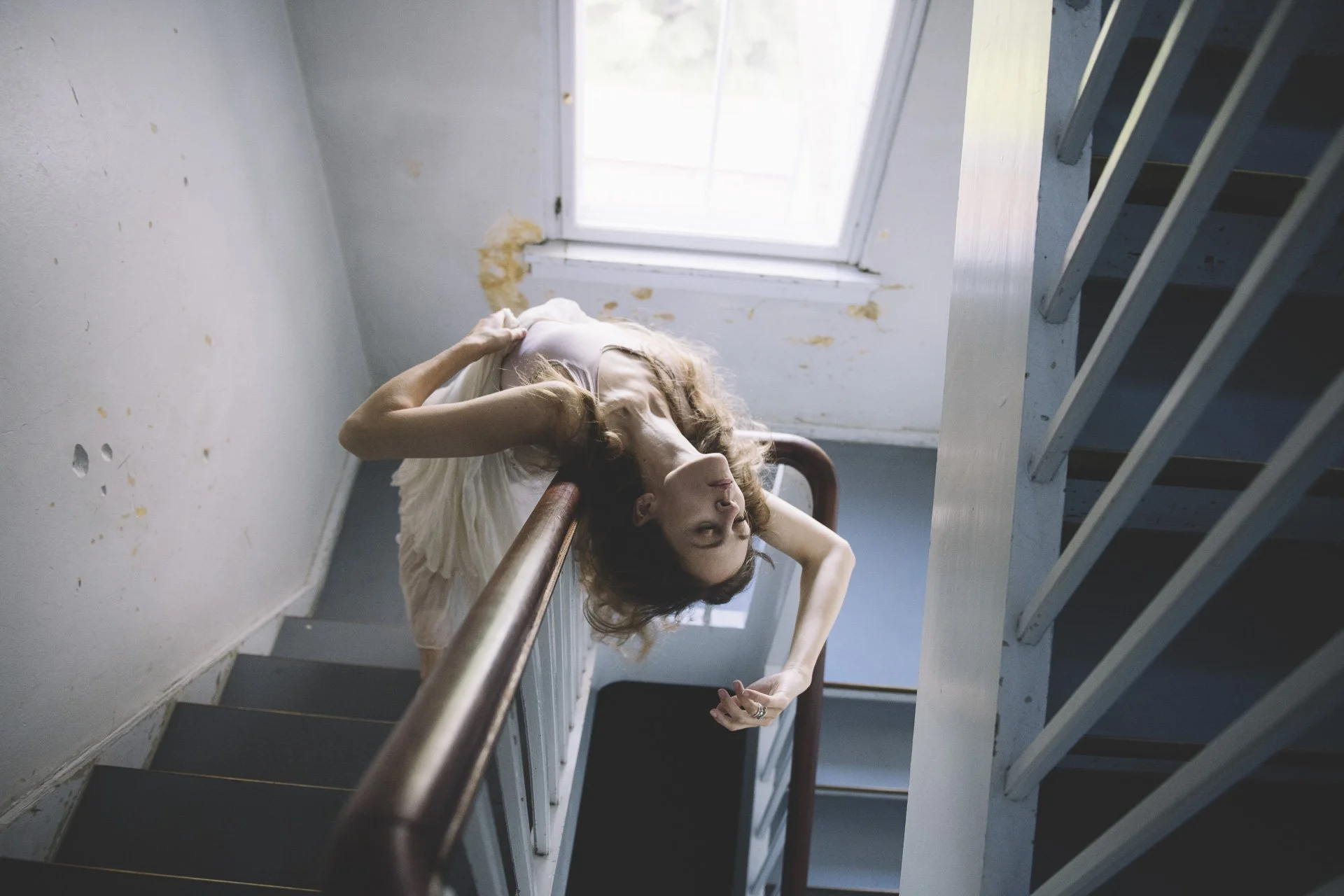 Eine Frau liegt auf der Treppe, lehnt am Geländer, vor einem Fenster mit natürlichem Licht, in einem alten Gebäude mit sichtbaren Schäden an der Wand.