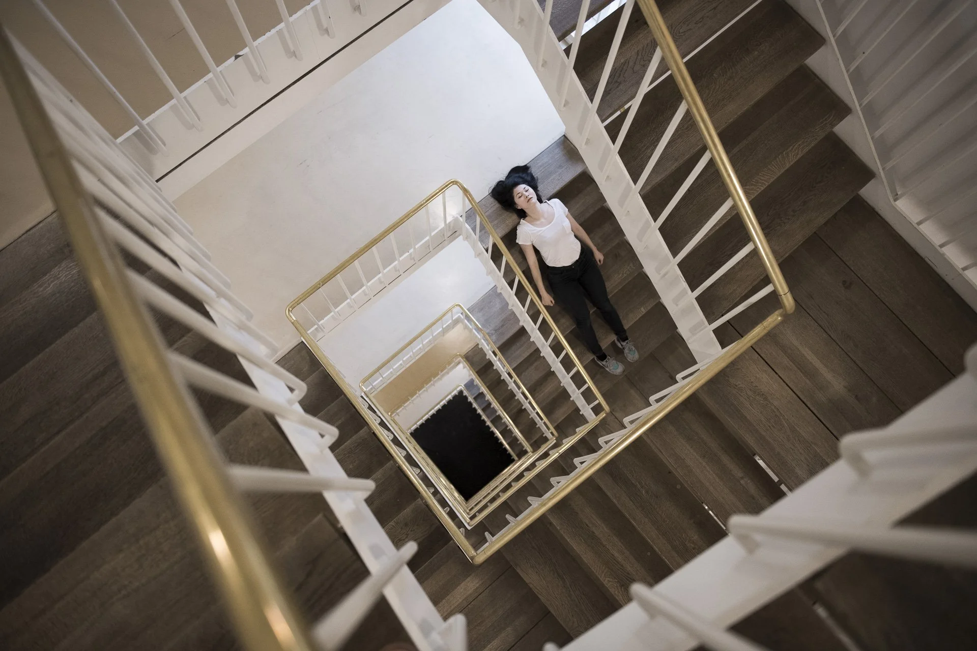 Eine Frau liegt in der Mitte einer Treppenhauswendeltreppe, die nach unten führt. Sie hat schwarze Haare, trägt eine weiße T-Shirt und schwarze Hosen, und liegt auf dem Boden der Treppenstufen. Die Treppe hat weiße Geländer mit goldfarbenen Handläufen, und die Treppenstufen sind aus dunklem Holz.