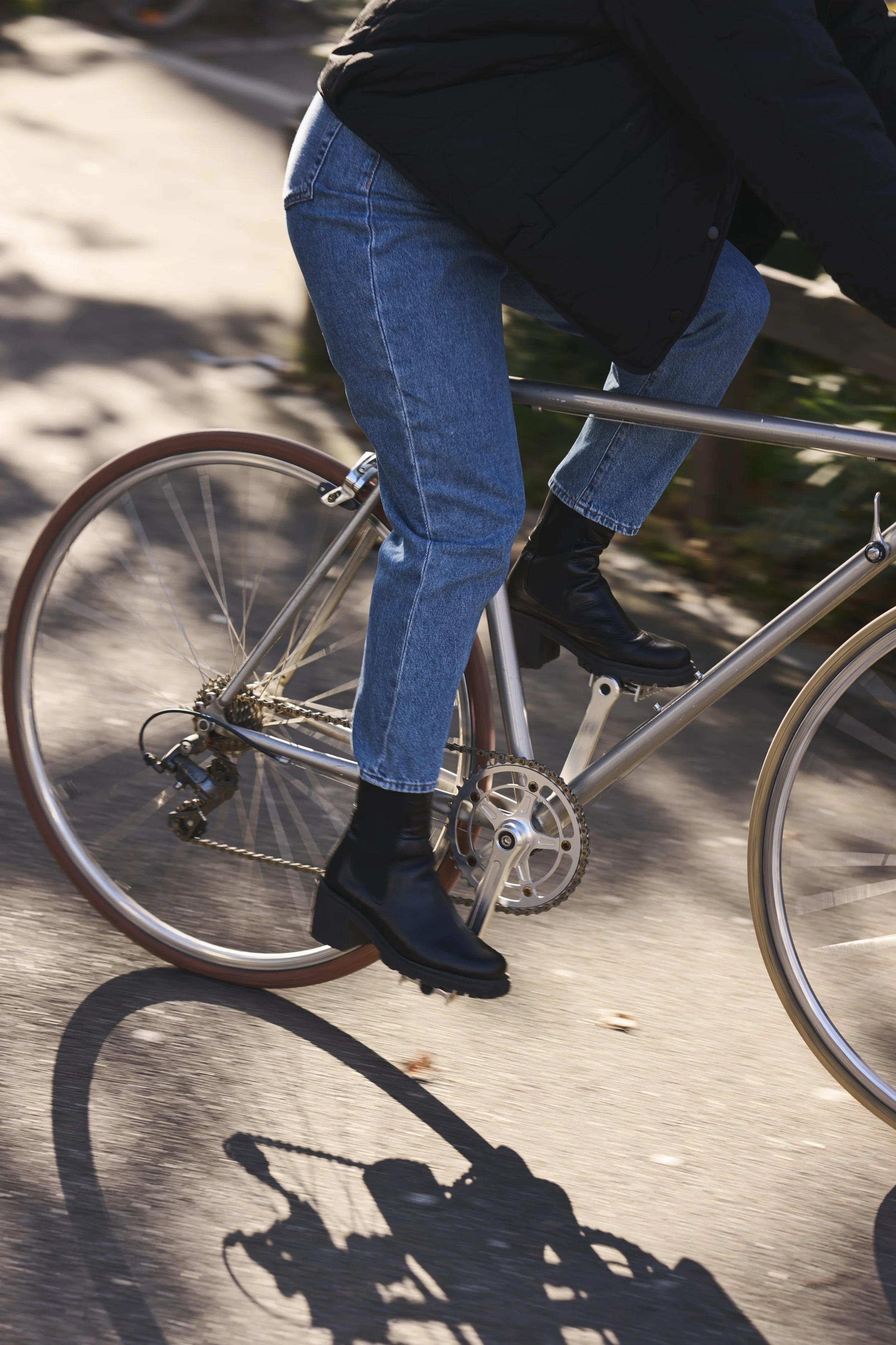 Nahaufnahme eines Fahrrads mit sichtbarem Pedal, Kette und Rahmen, während eine Person in blauen Jeans und schwarzen Schuhen darauf fährt. Der Schatten des Fahrrads ist auf der Straße sichtbar, während die Hintergründe unscharf sind.