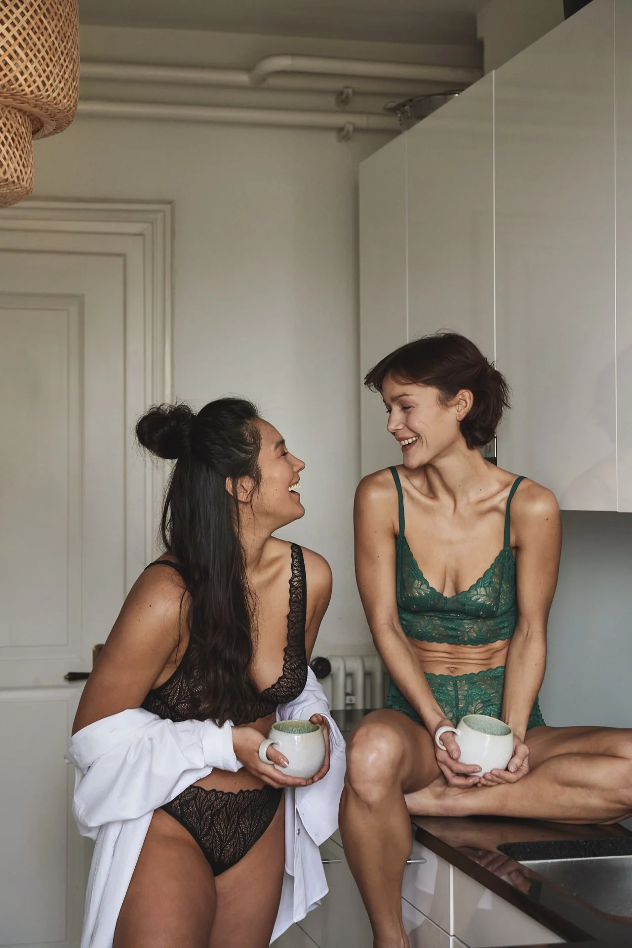 Zwei Frauen in sexy Unterwäsche und Männer in Unterhosen unterhalten sich in einer modernen Küche, lachen und halten Tassen.