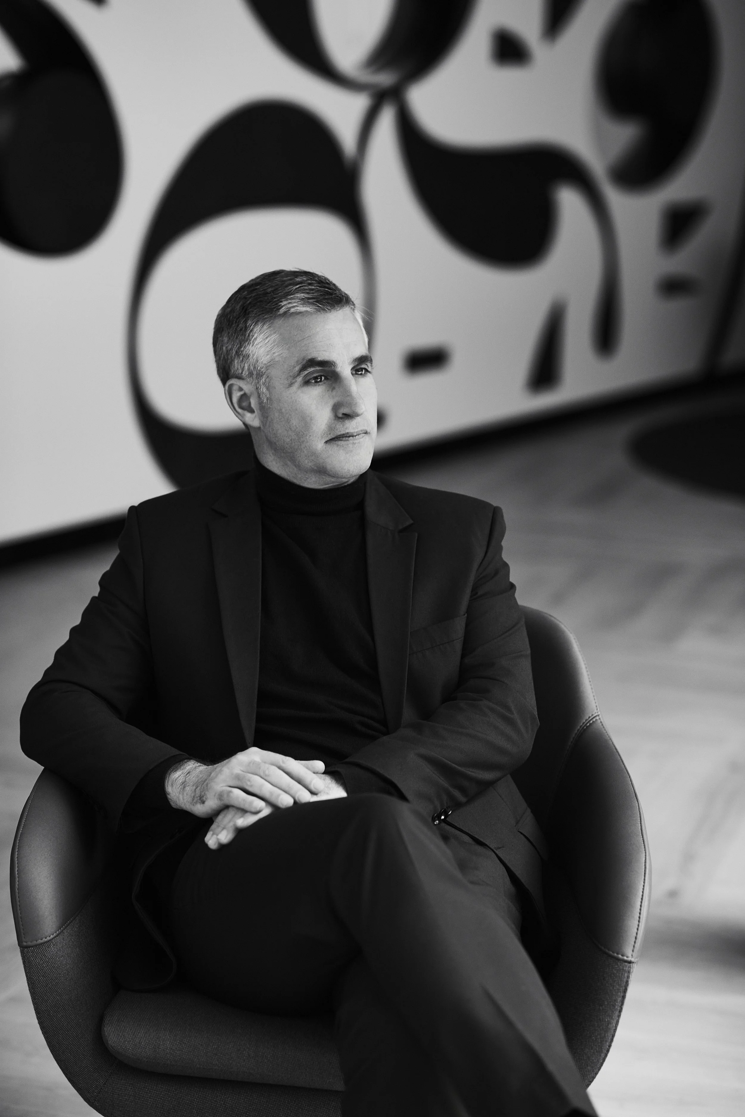 Schwarz-weiß-Fotografie eines Mannes sitzt nachdenklich in einem modernen Stuhl vor einem großen Kunstwerk mit abstrakten schwarzen Formen auf weißem Hintergrund.