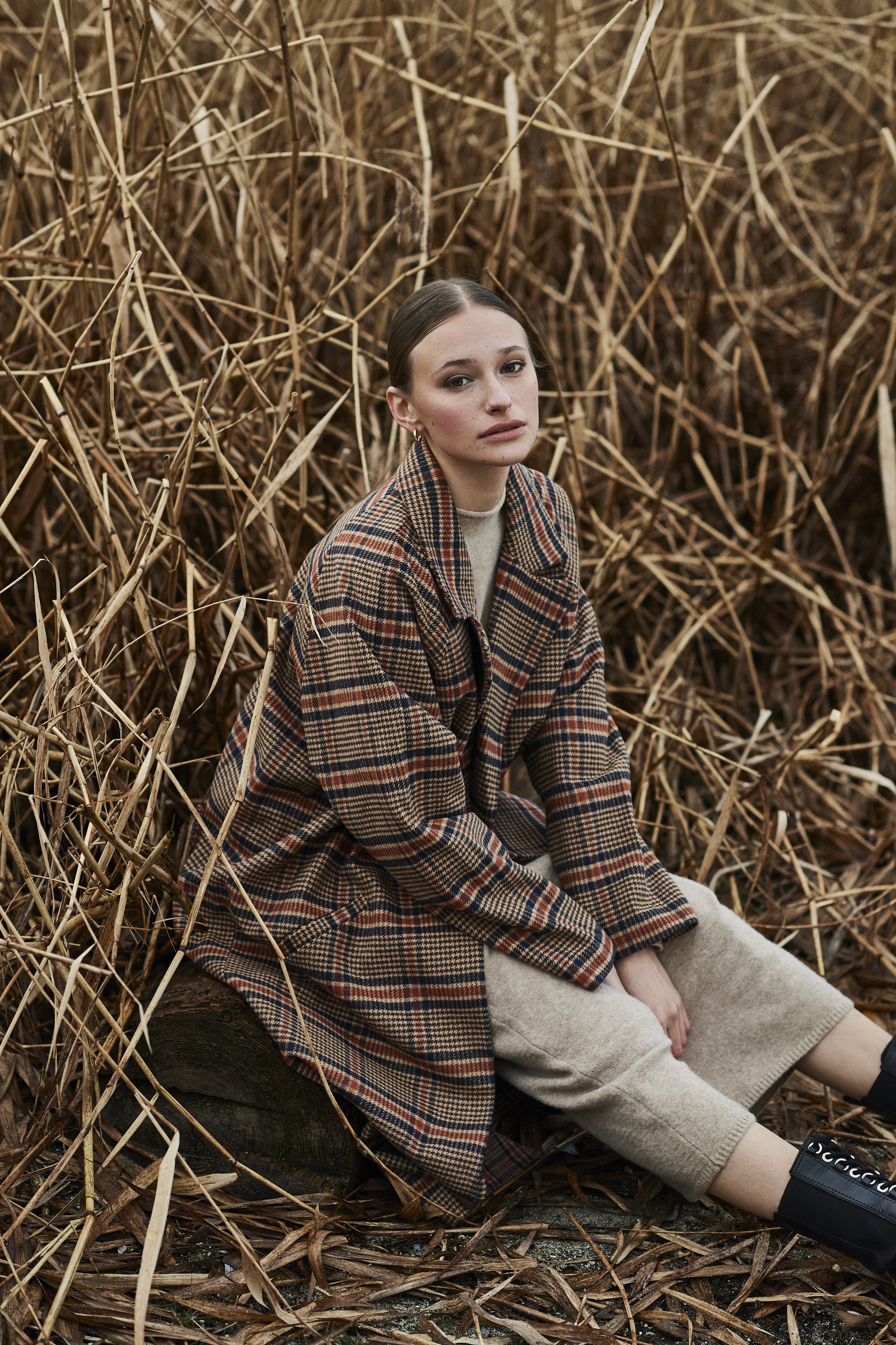 Junge Frau sitzt im Laub einer abgestorbenen Pflanze, trägt ein kariertes Mäntel, beige Hosen und schwarze Turnschuhe.