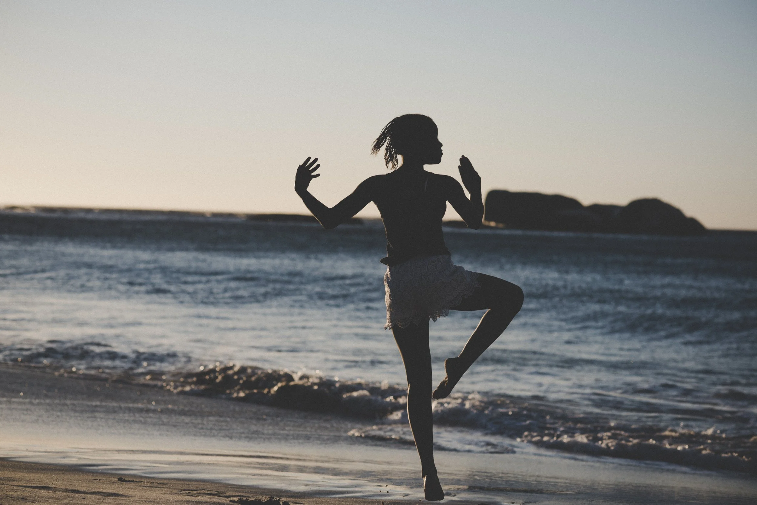 Silhouette einer Frau beim Yoga am Strand bei Sonnenuntergang, mit Wasser und Felsen im Hintergrund.