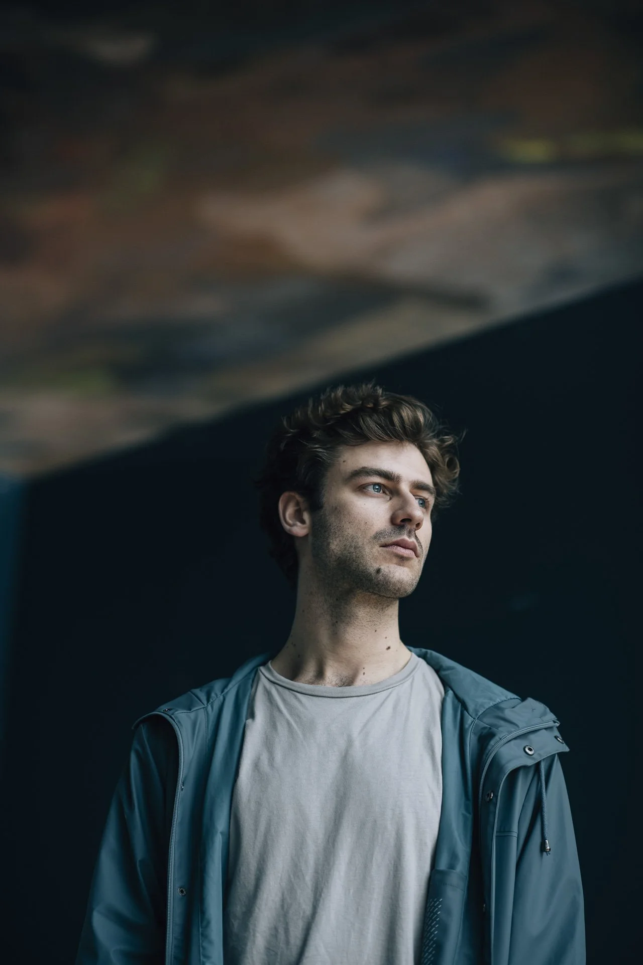 Junger Mann mit braunen, lockigen Haaren, blauen Augen und leichter Barthaare steht vor einem dunklen Hintergrund, trägt ein graues T-Shirt und eine blaue Jacke.