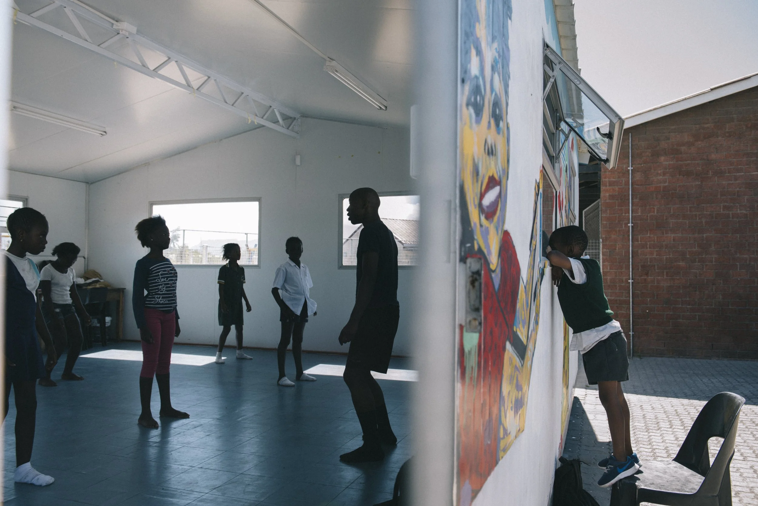 Junge und Mädchen in einem Turnraum, einige stehen, einer lehnt an einer Wand, andere sind in einer Linie, eine Person steht gegenüber, innerhalb eines Schulgebäudes mit großen Fenstern und Deckenbeleuchtung, draußen sieht man eine Brücke und einen Himmel.