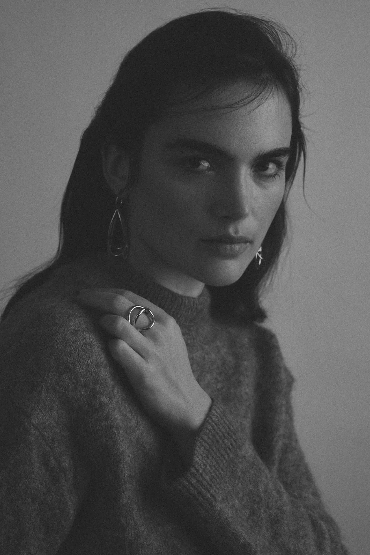 Schwarz-Weiß-Fotografie eines jungen Mädchens mit dunklen Haaren, das direkter Blick in die Kamera zeigt, mit einer Hand auf ihrer Schulter und trägt Ohrringe und Ringe, trägt einen Pullover.