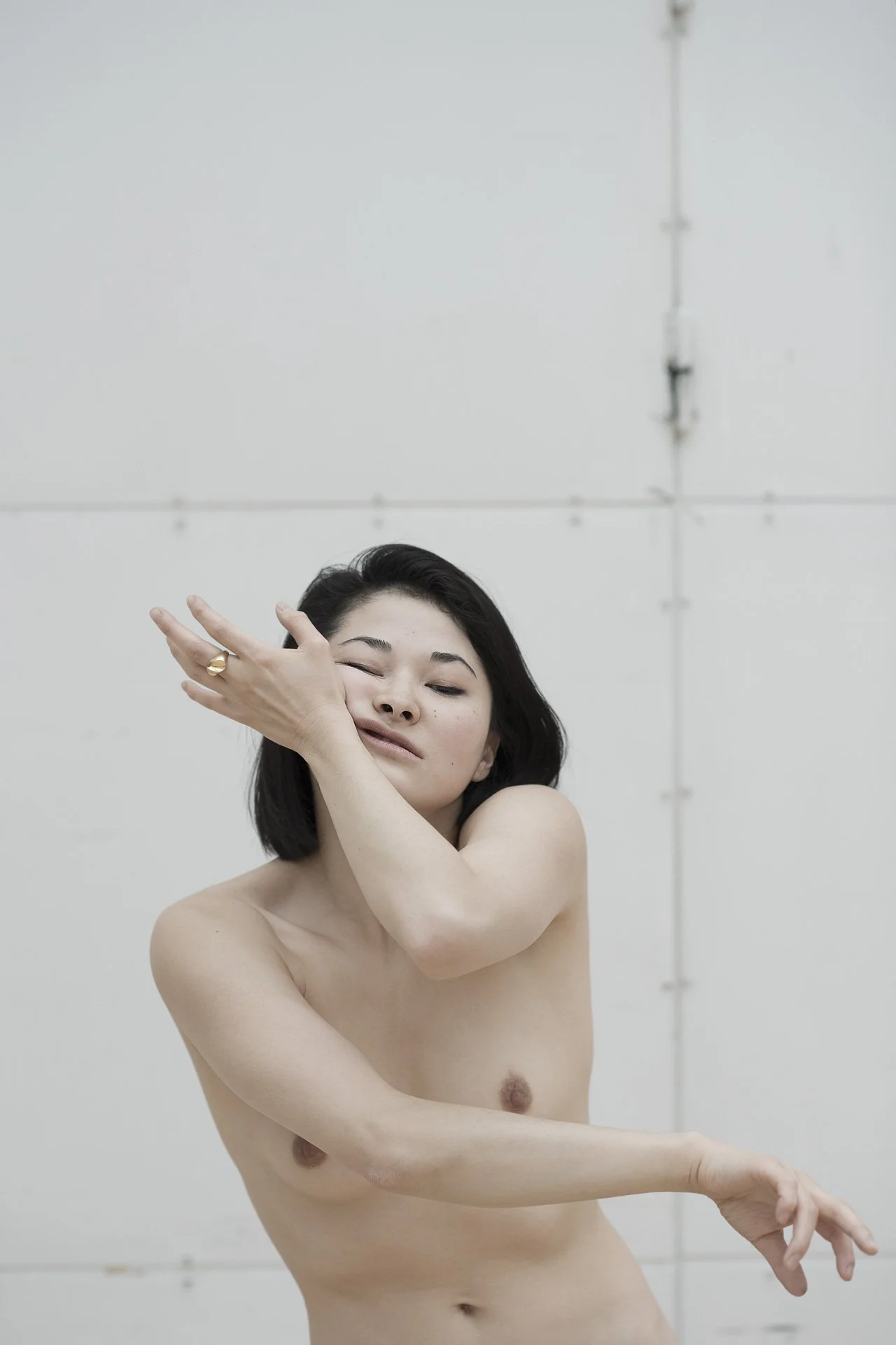 Junge Frau ohne Kleidung posiert vor hellem Hintergrund, mit geschlossenen Augen und einer Hand am Gesicht, lächelnd