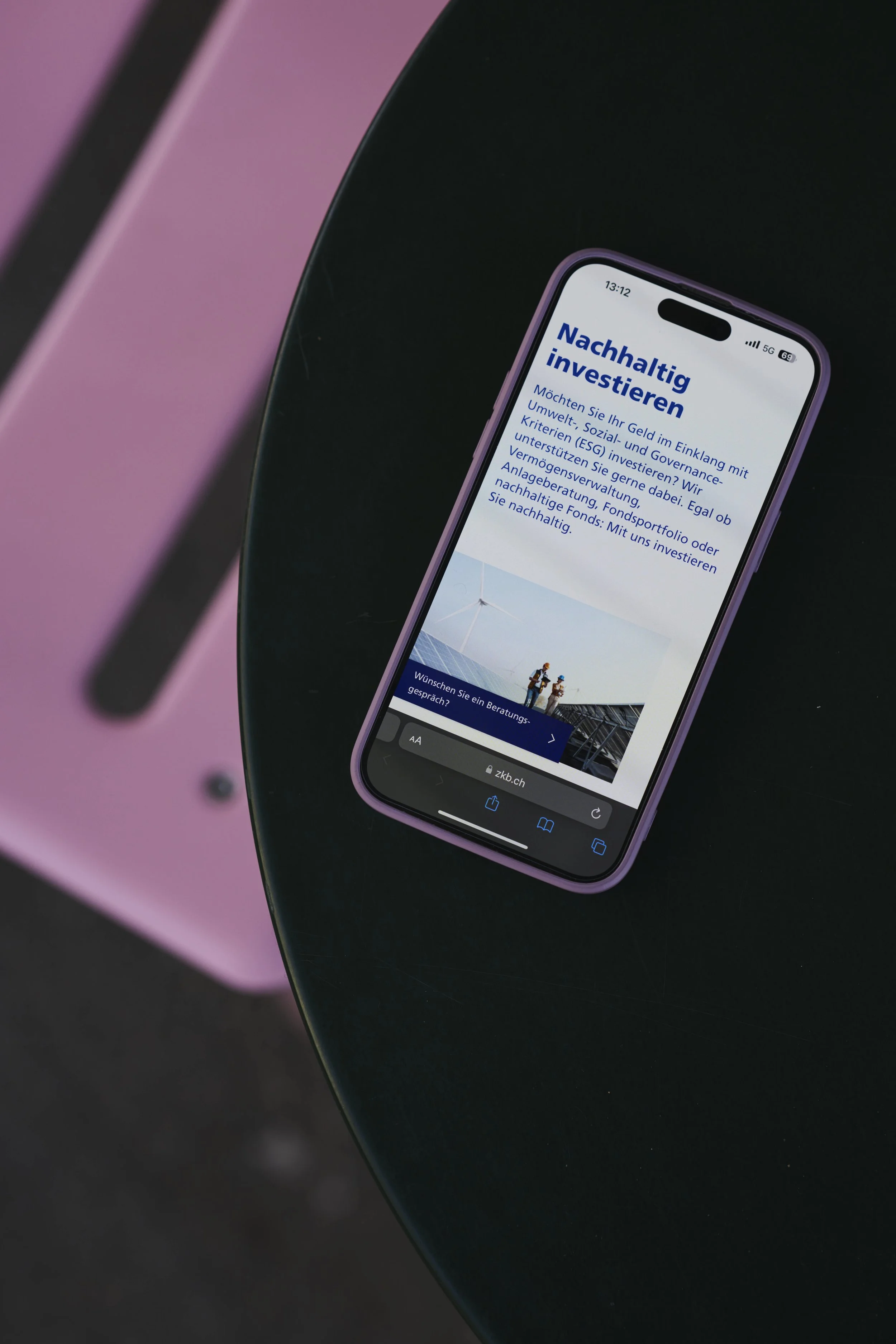 Nahaufnahme eines Smartphones, das eine Webseite über nachhaltige Investitionen zeigt. Das Handy liegt auf einer schwarzen Oberfläche, im Hintergrund ist eine pinkfarbene Fläche sichtbar.